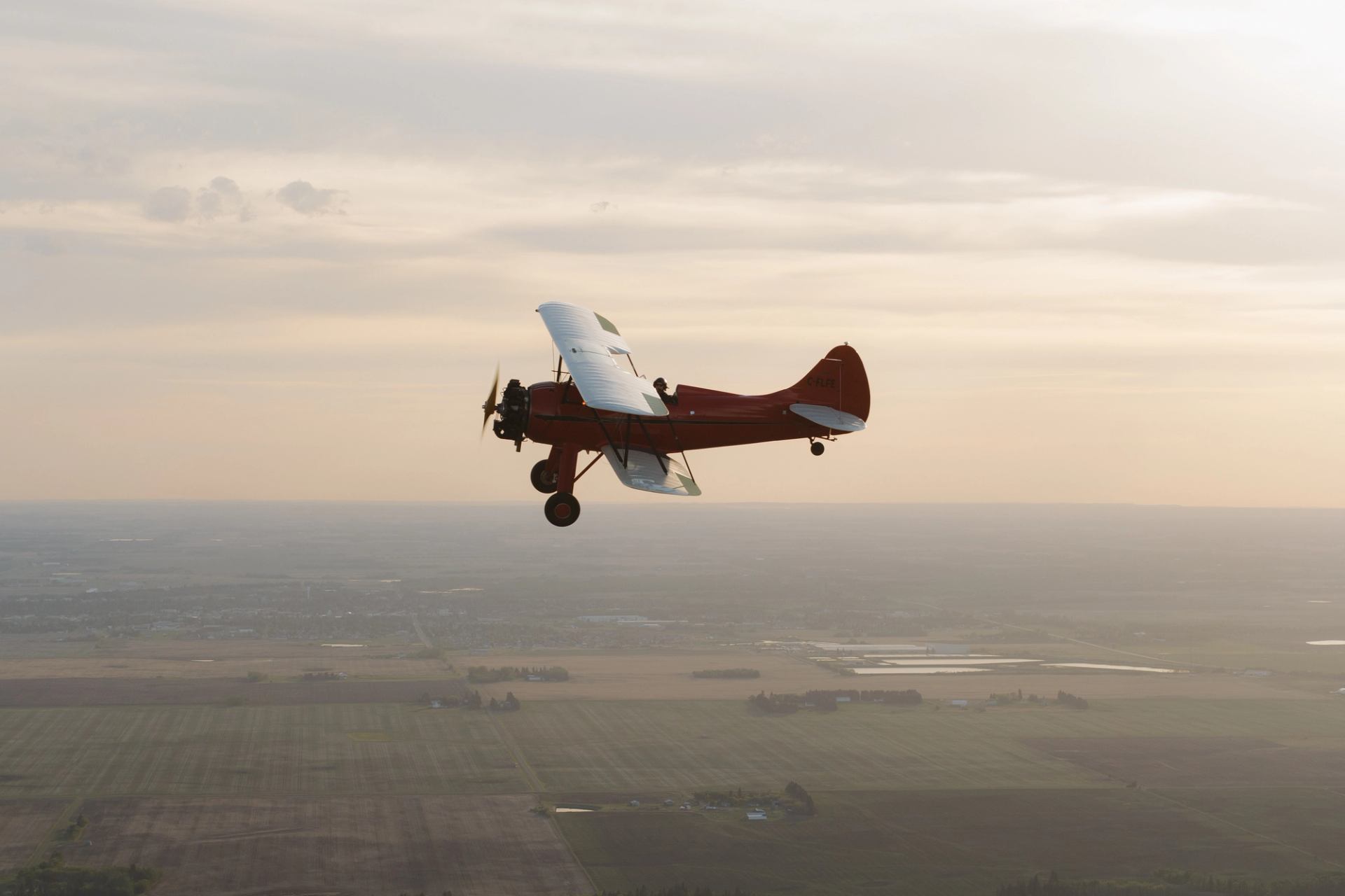 Red biplane flies above flat farmland landscape under hazy, pale sky.