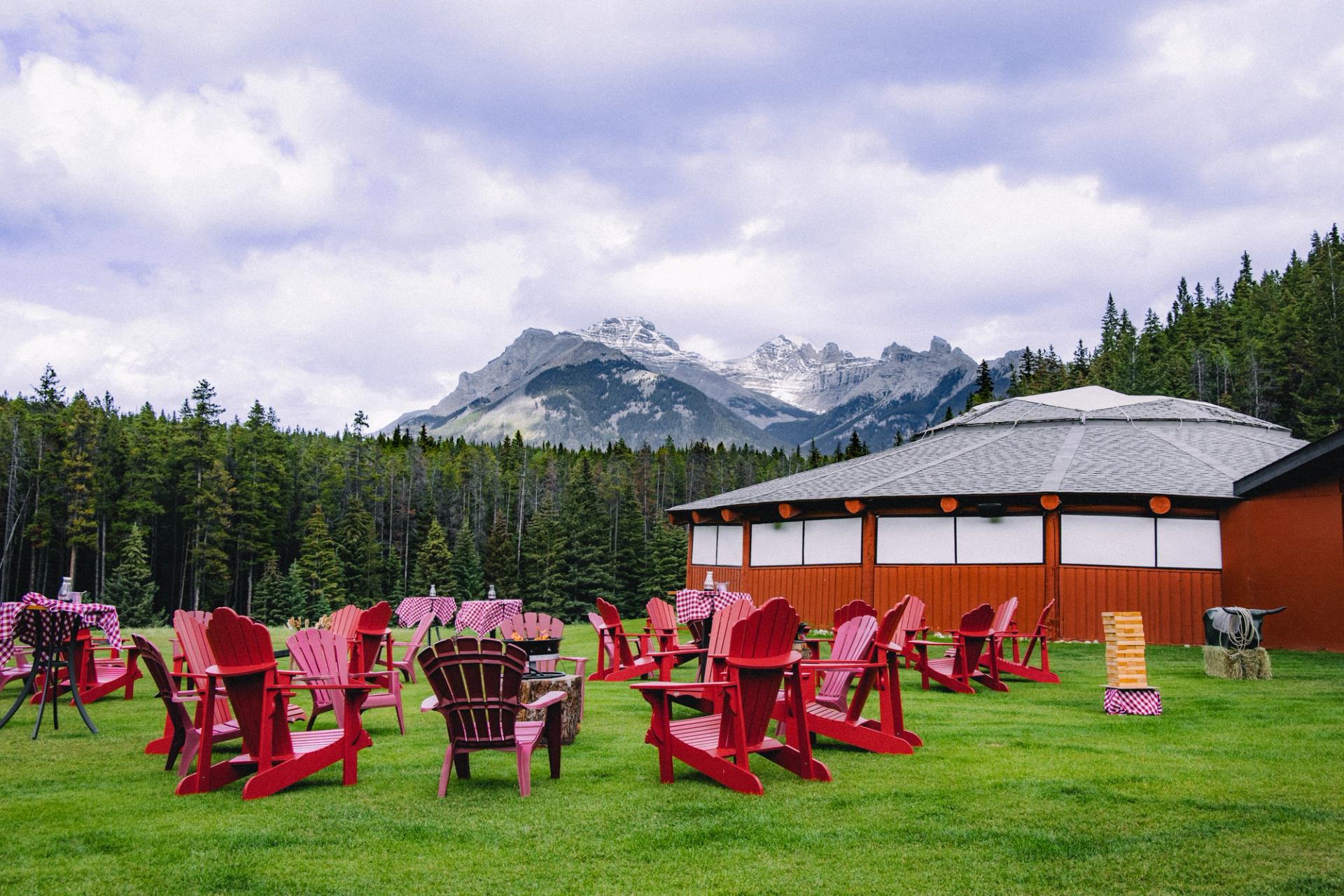 Red Adirondack chairs on a grassy lawn in front of a red building, with a forest and snow-capped mountains in the background under a cloudy sky.