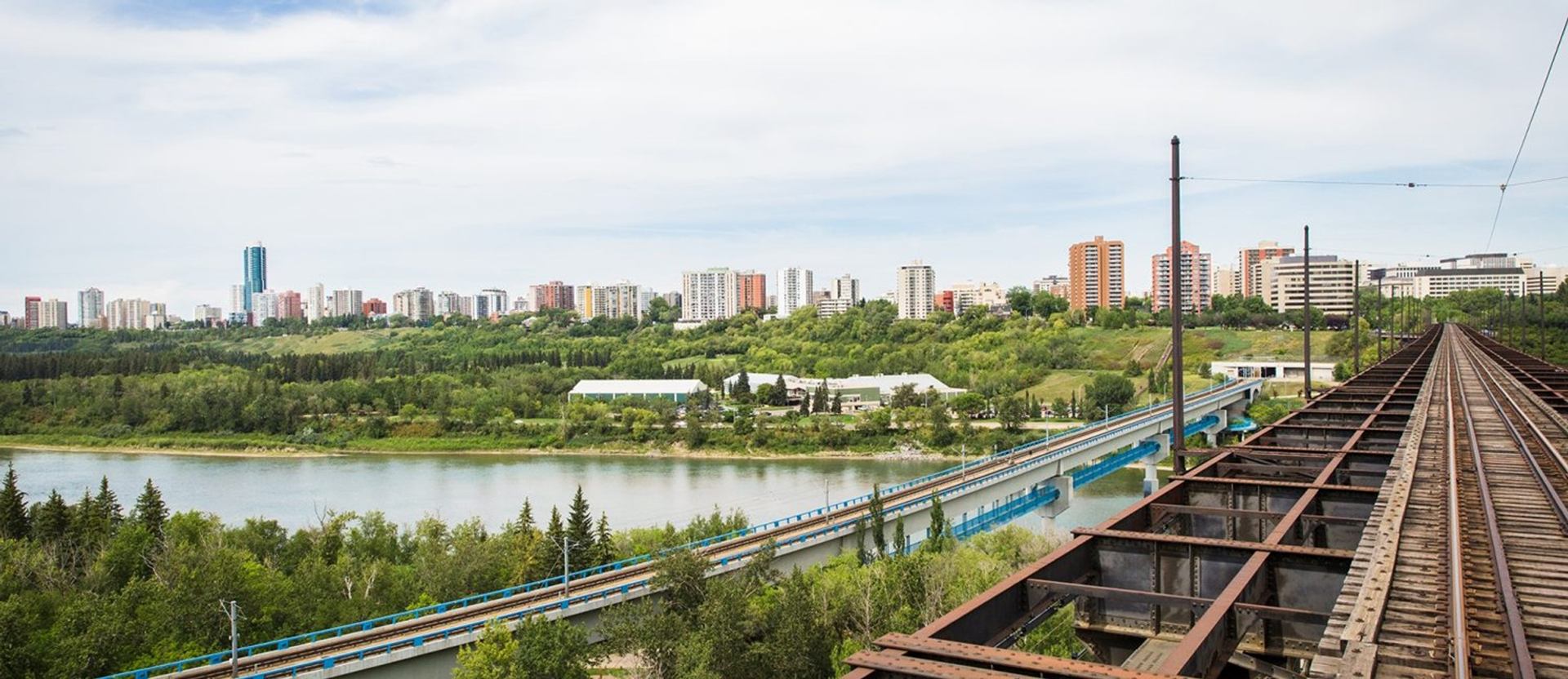 River valley with bridges, railway tracks and distant city skyline under cloudy sky.