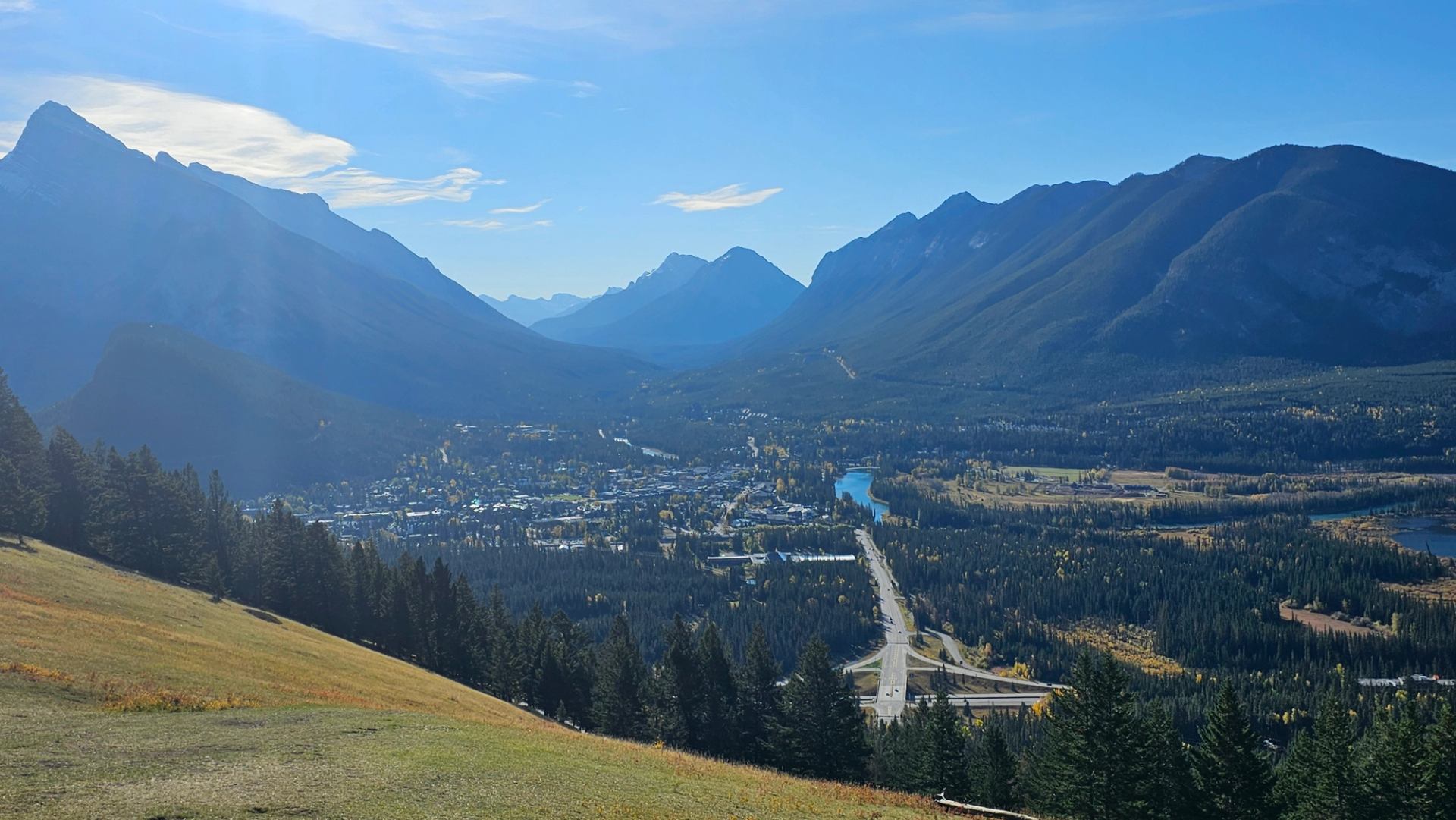 Mountain valley with town, highway, river and forests viewed from grassy hillside.