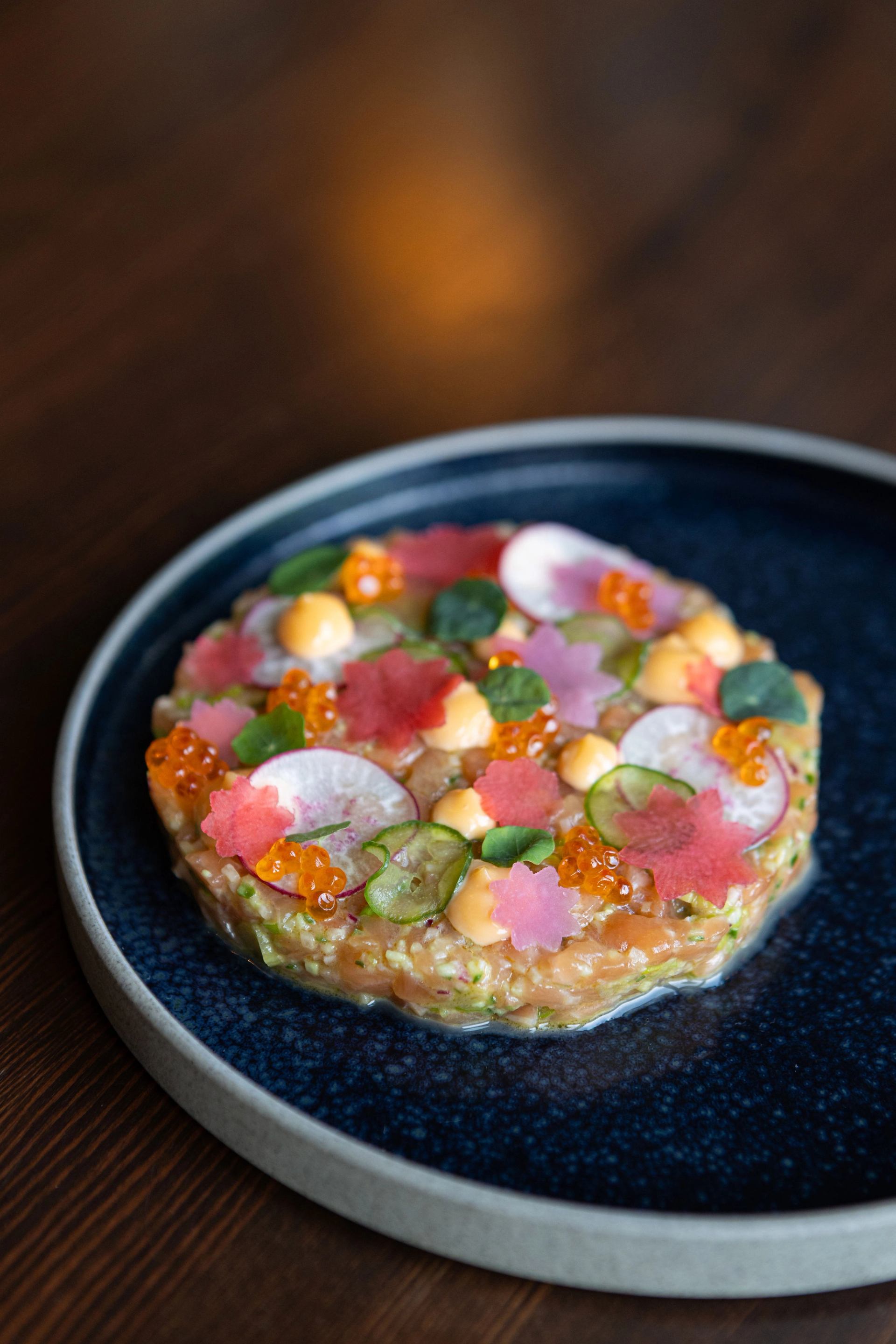 Salmon tartare topped with radish, cucumber, roe, greens and flower-shaped garnishes.