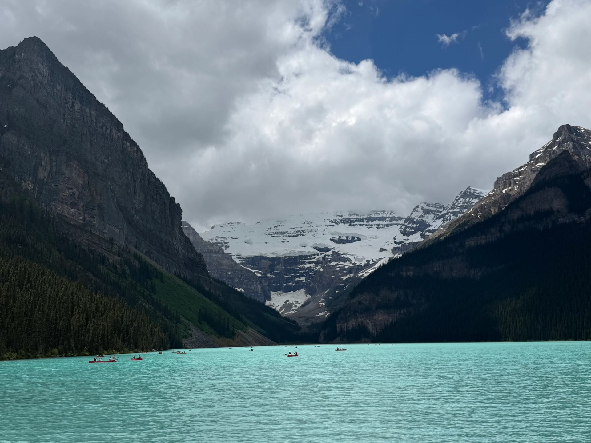Turquoise mountain lake with small canoes, steep forested slopes and snowy peaks under clouds.