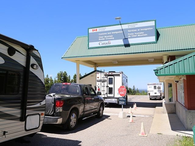 Border checkpoint with vehicles, trailer, and bilingual Canada customs sign.