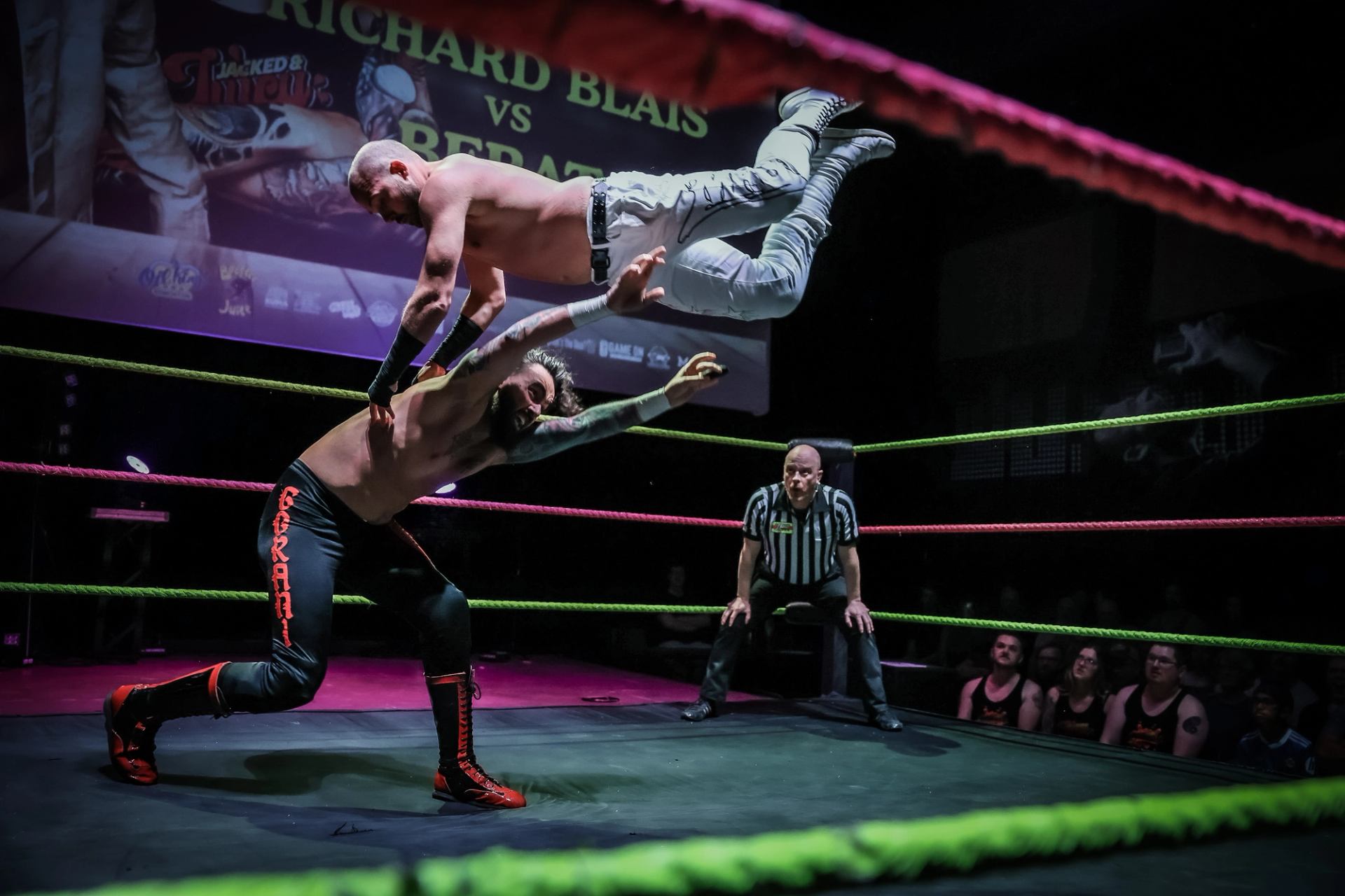 Professional wrestler performs flying move onto opponent in ring as referee watches closely.