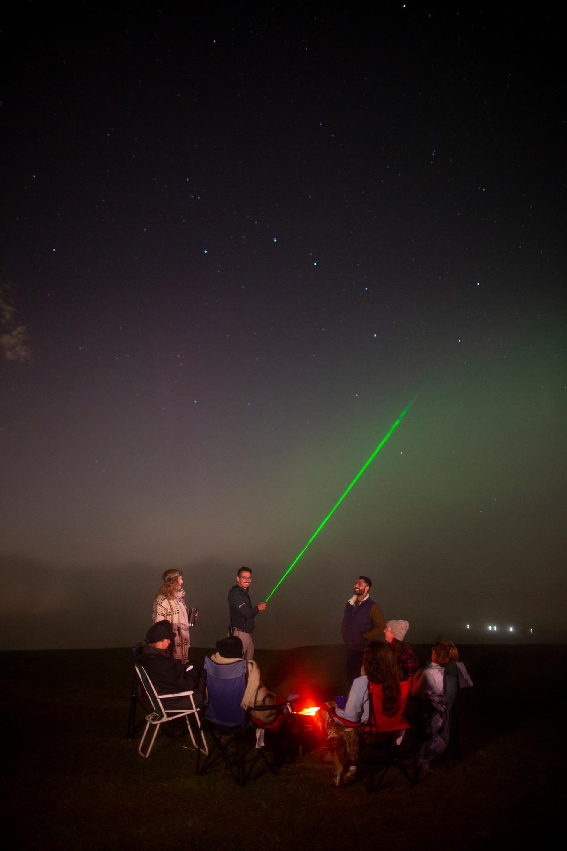 Group gathers around a campfire as a green laser points into the starry sky.