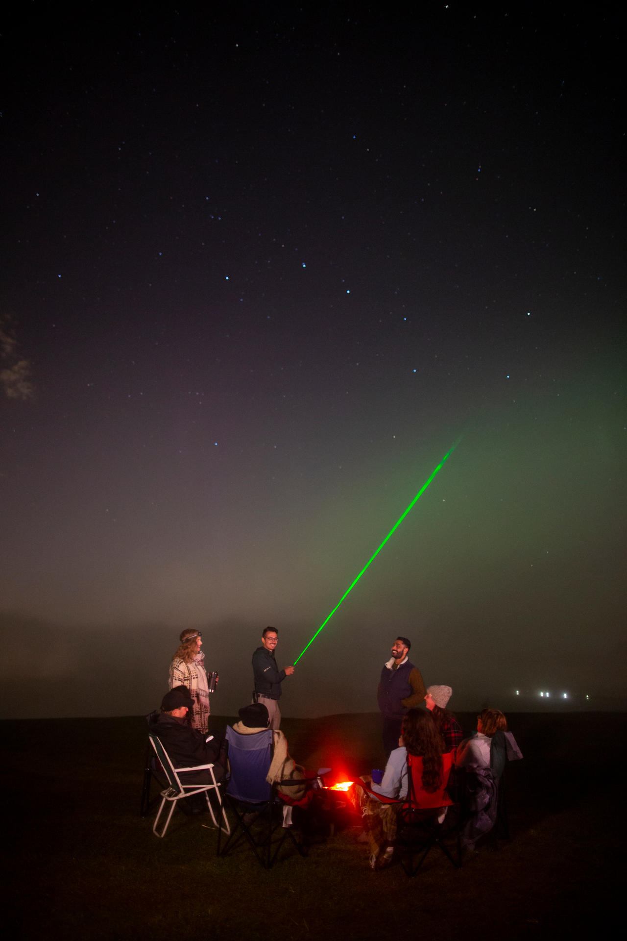 Group gathers around a campfire as a green laser points into the starry sky.