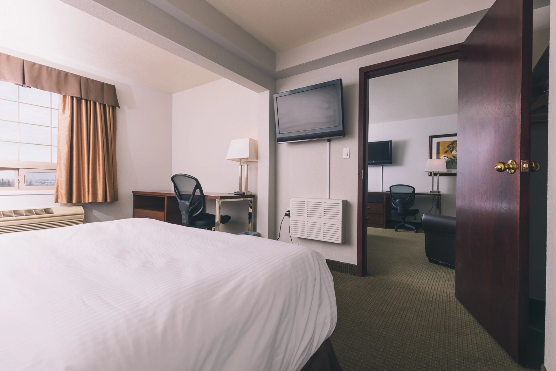 Hotel room with bed, desk, chair, lamp, wall-mounted TV and adjoining room.
