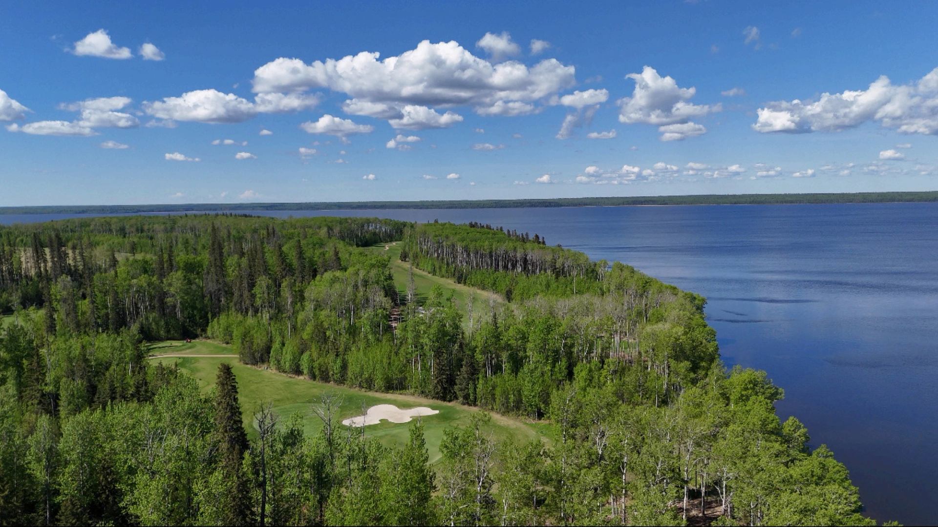 Forested golf course beside a large blue lake under scattered clouds.