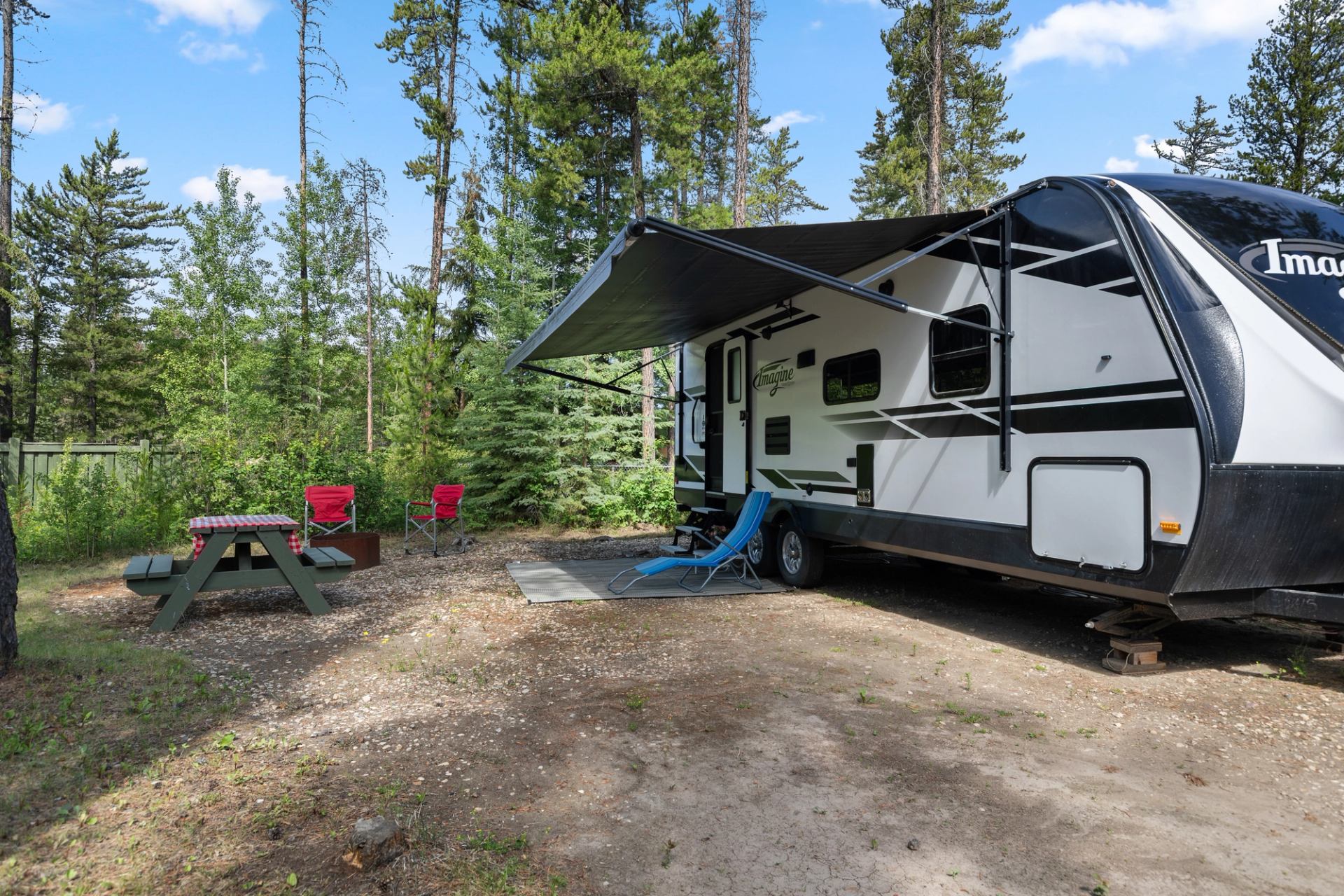 Travel trailer parked at a wooded campsite with chairs, picnic table, and shaded outdoor space.