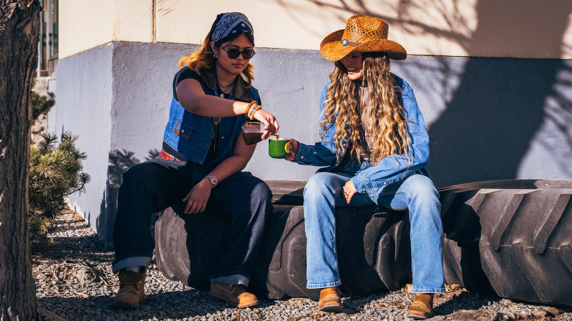 Two people in western outfits sitting outdoors on large tires, sharing a green coffee mug.