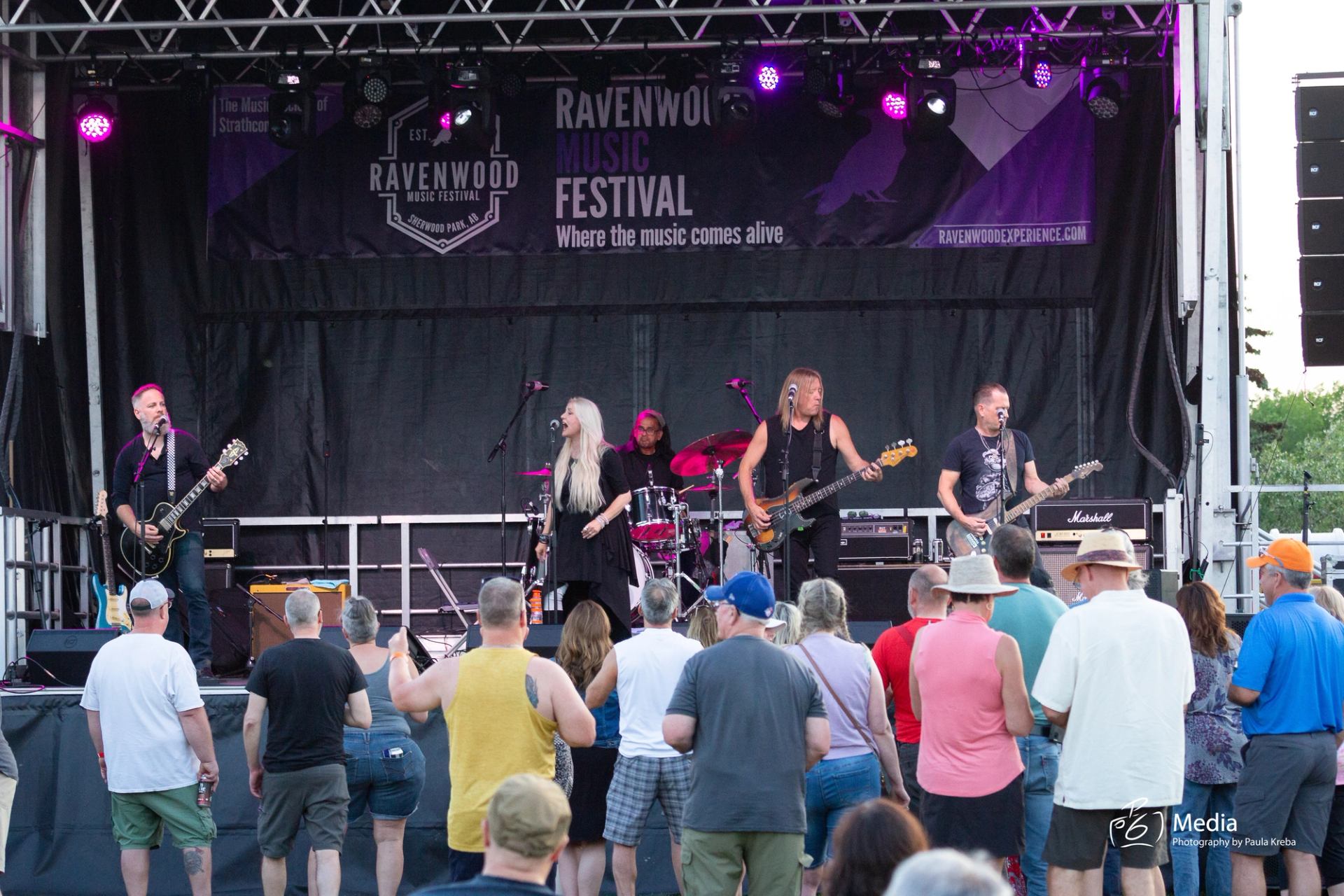 Audience seated on grass watching a live performance on an outdoor stage.