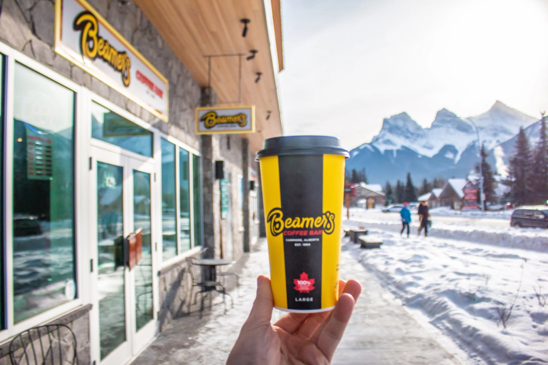 Hand holding a Beamer’s Coffee Bar cup outside the café with snowy mountains in the background.