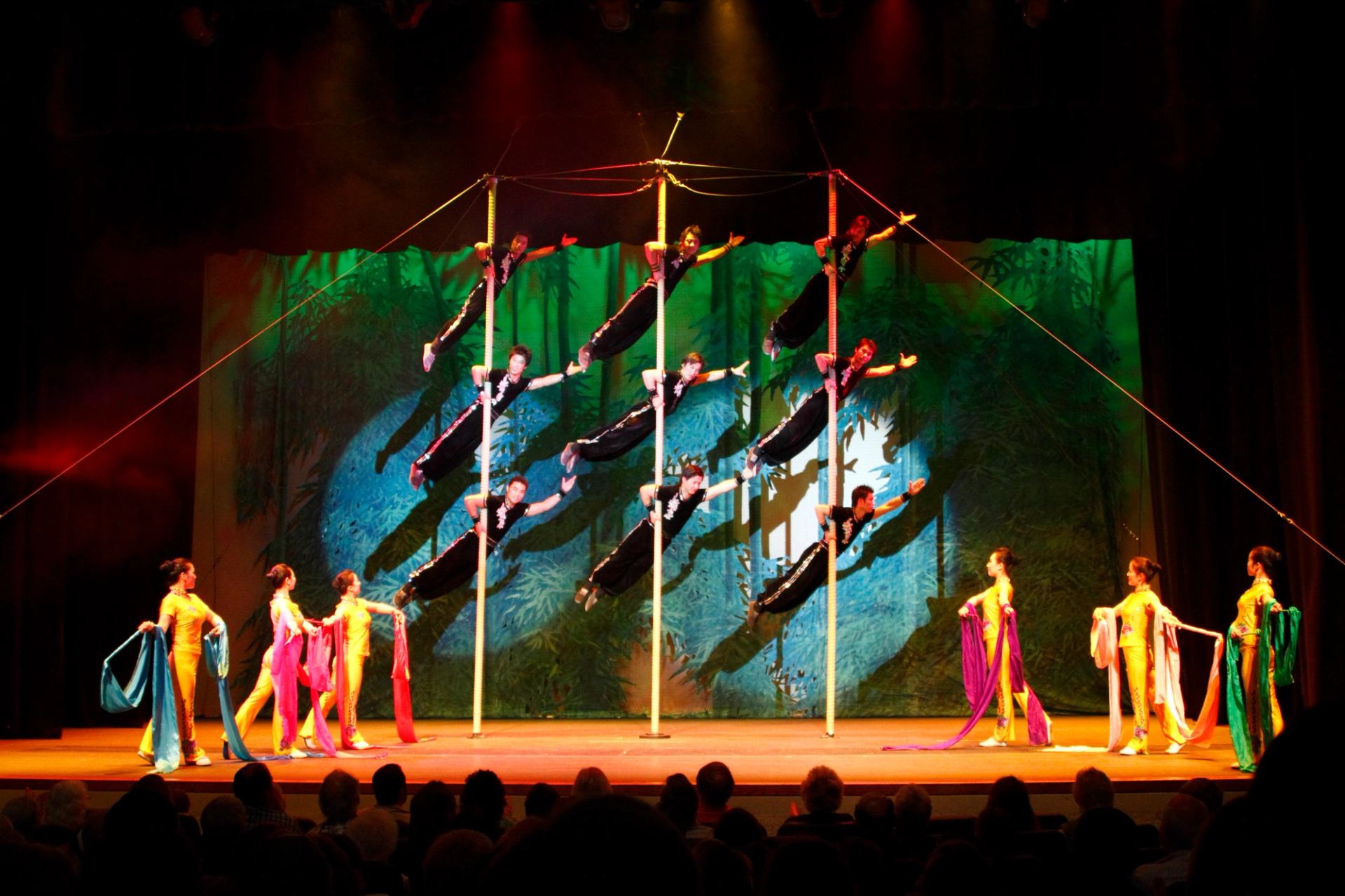 Acrobats balance horizontally on tall poles as dancers hold colourful flowing fabric onstage.