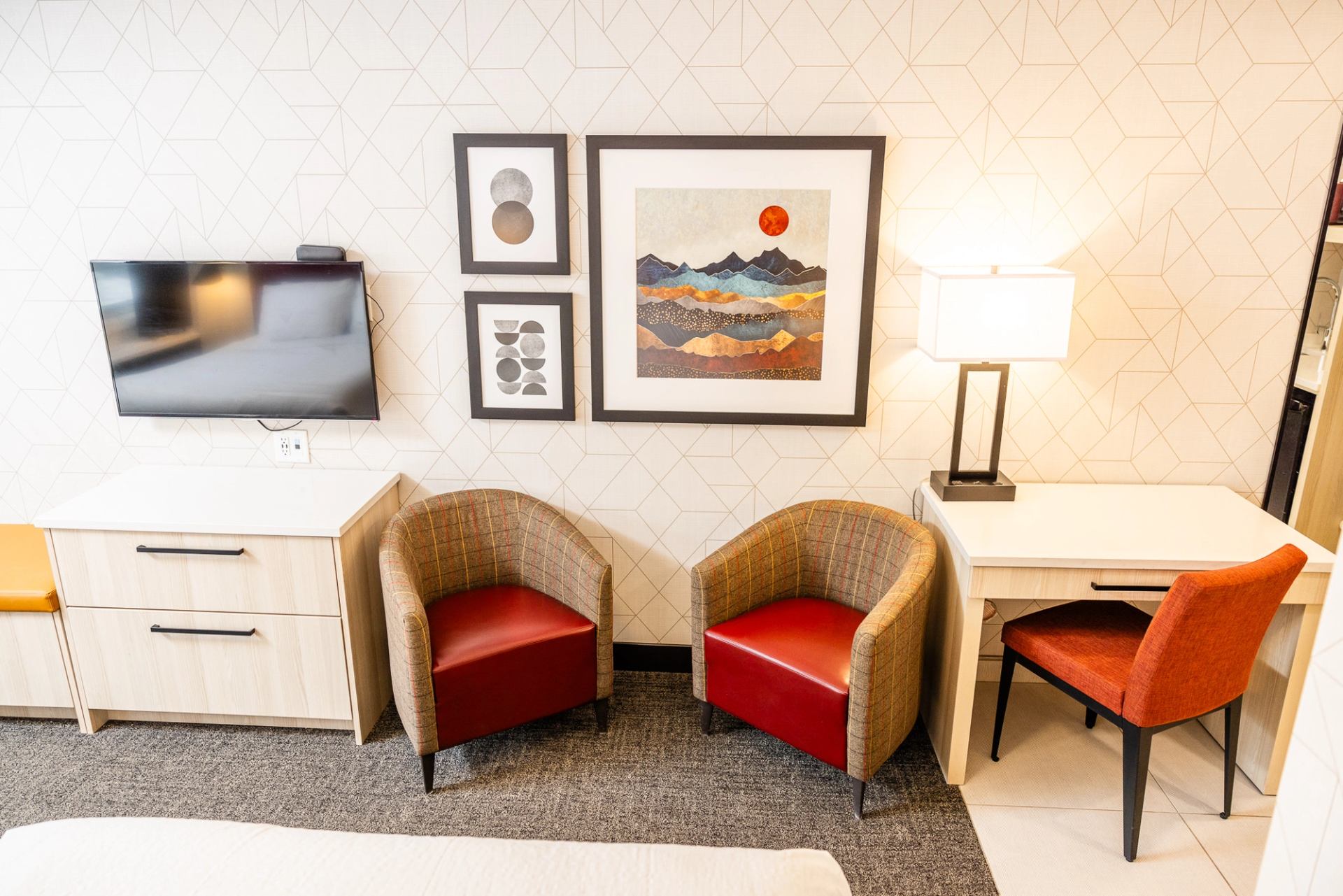 Modern hotel room interior featuring two red armchairs, a white desk, and mountain-themed wall art.