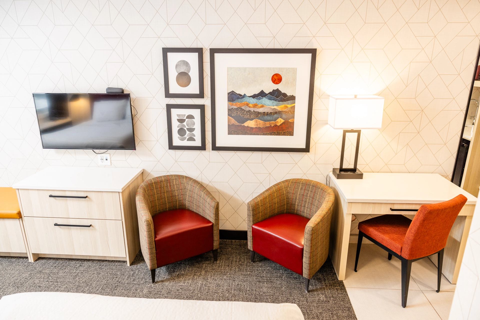 Modern hotel room interior featuring two red armchairs, a white desk, and mountain-themed wall art.