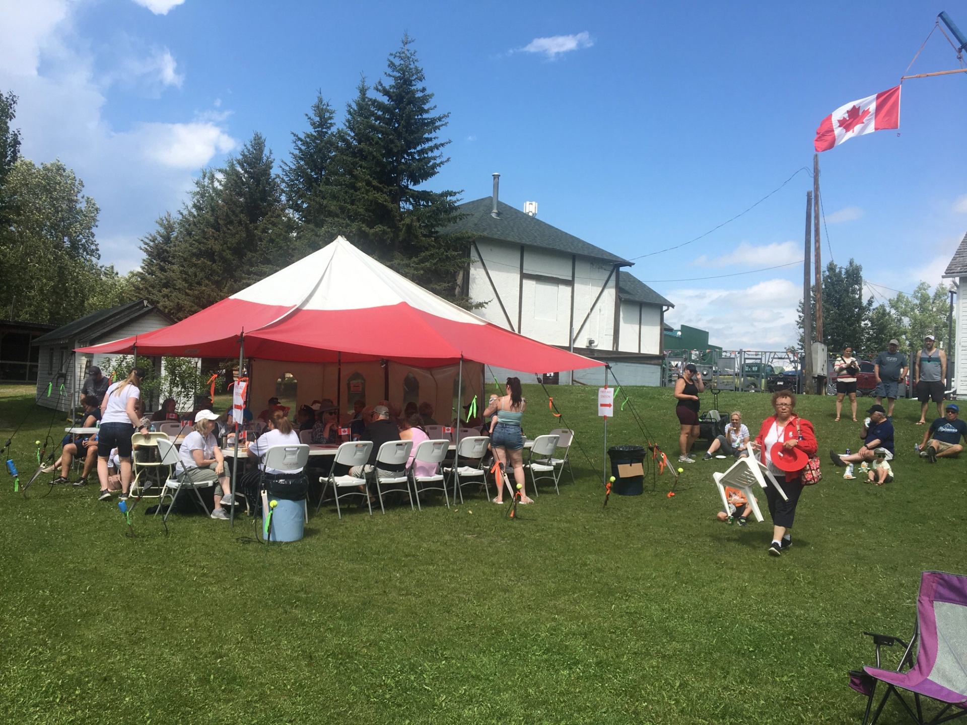 Outdoor gathering under a red and white tent with people seated on lawn chairs.
