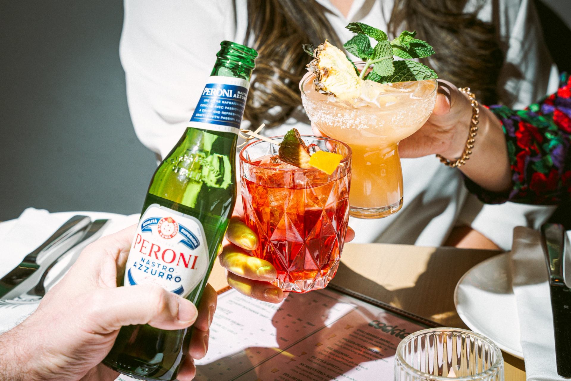 Hands raising cocktails and a Peroni bottle for a toast at a restaurant table.
