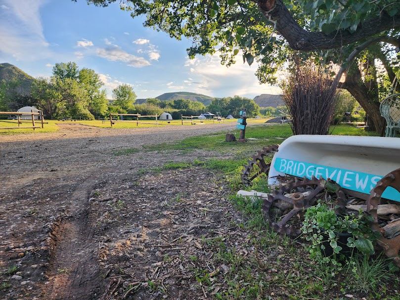 Campground with yurts, gravel road, trees, hills, and Bridgeviews sign on old boat.