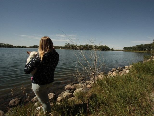 A lady stood on the shore with a dog in her arms looking at the lake.