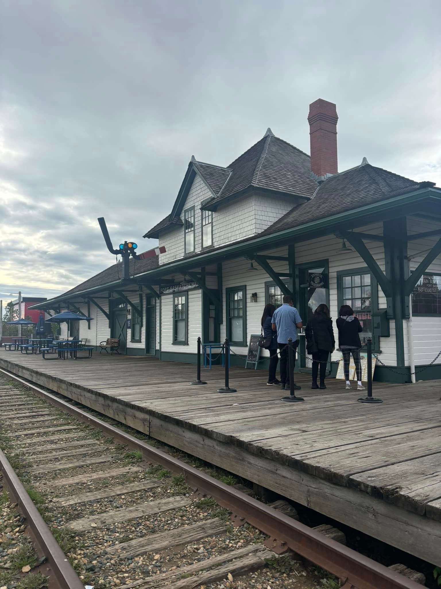 Historic train station building with a wooden platform, railroad tracks, and a few people.
