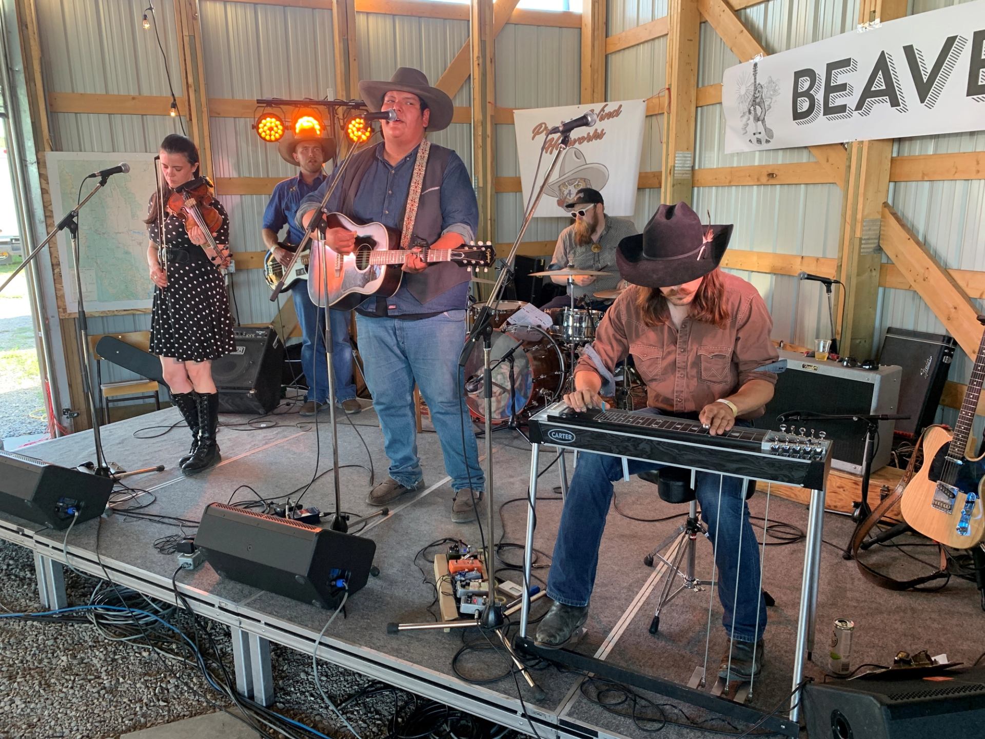 A country band performs on stage with a fiddler, singer-guitarist, pedal steel player, and drummer; several members wear cowboy hats.