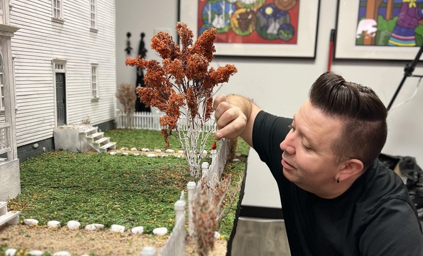 Person holds a small tree display outdoors in front of a building and framed artwork.