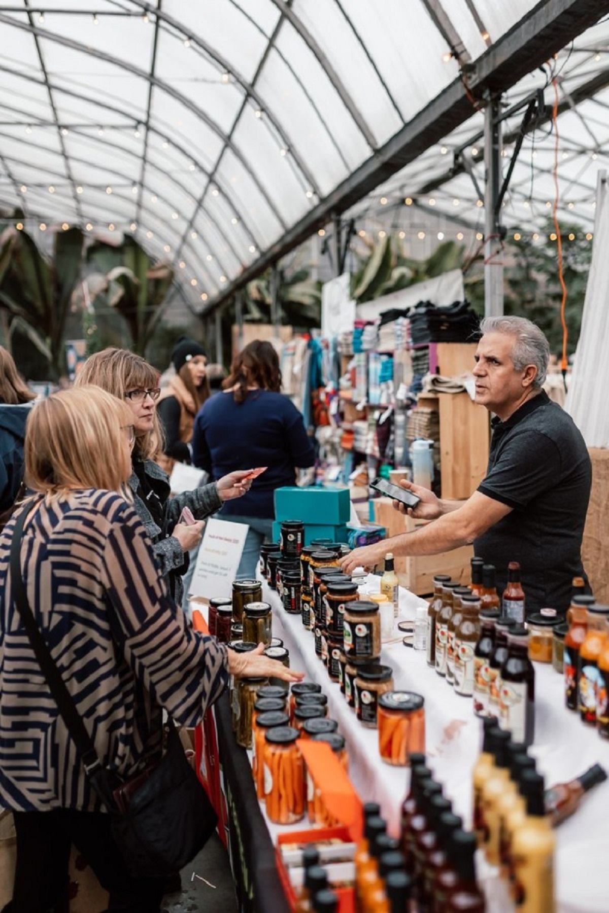 Shoppers browsing vendor stall in festive greenhouse market setting.
