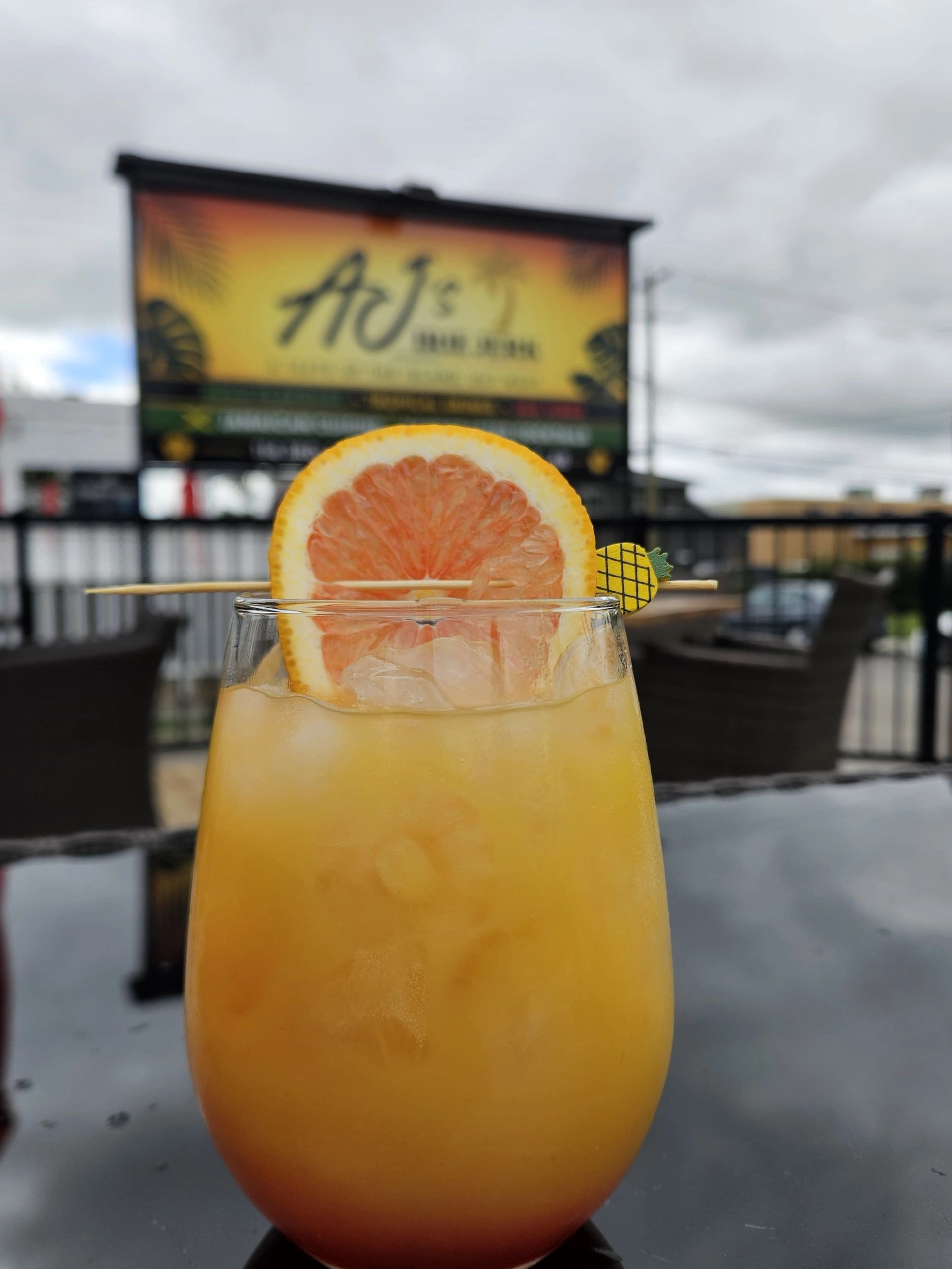 Orange iced drink with grapefruit garnish sits before a blurred AJ's sign.
