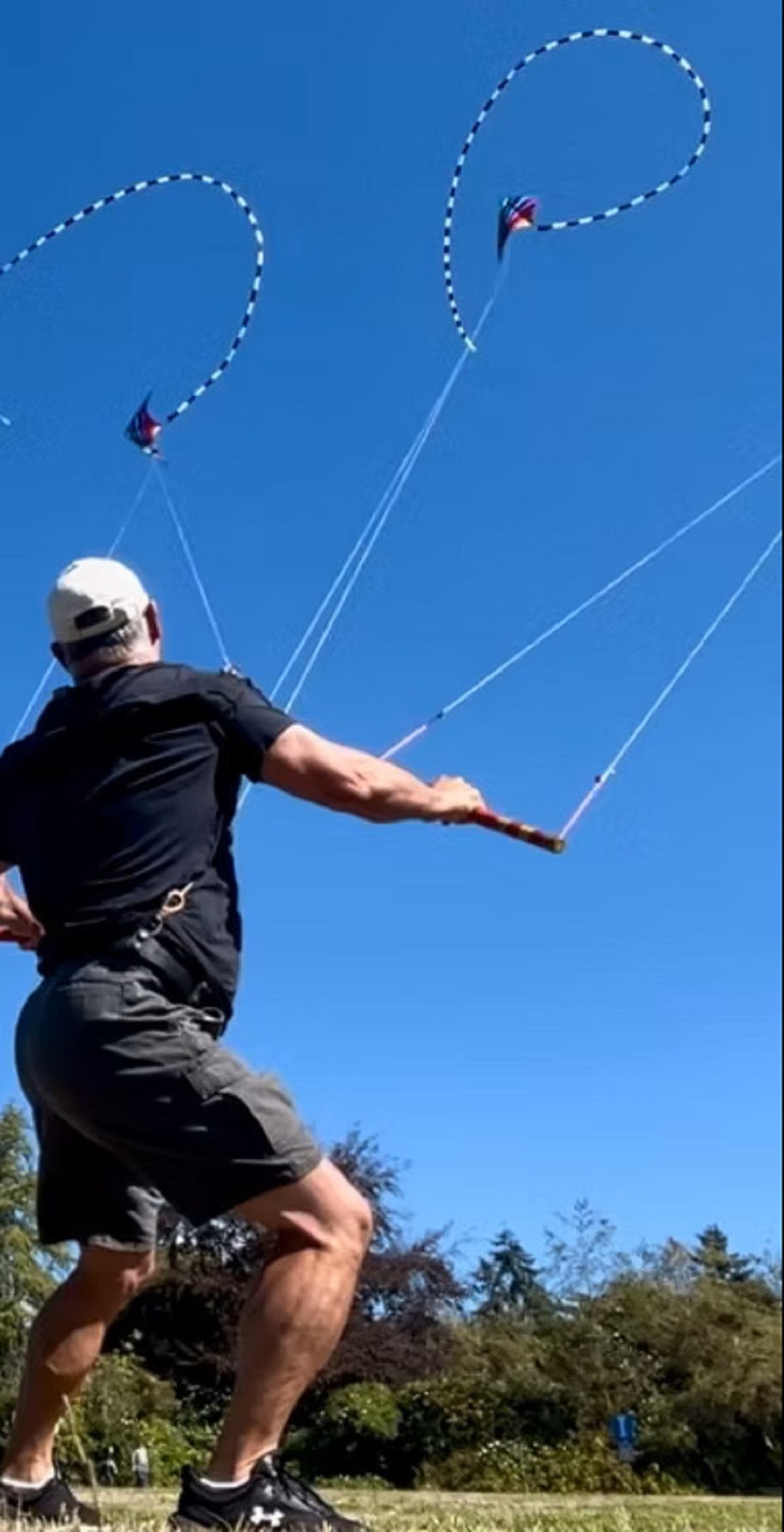Person flying two stunt kites in a park under a clear blue sky.
