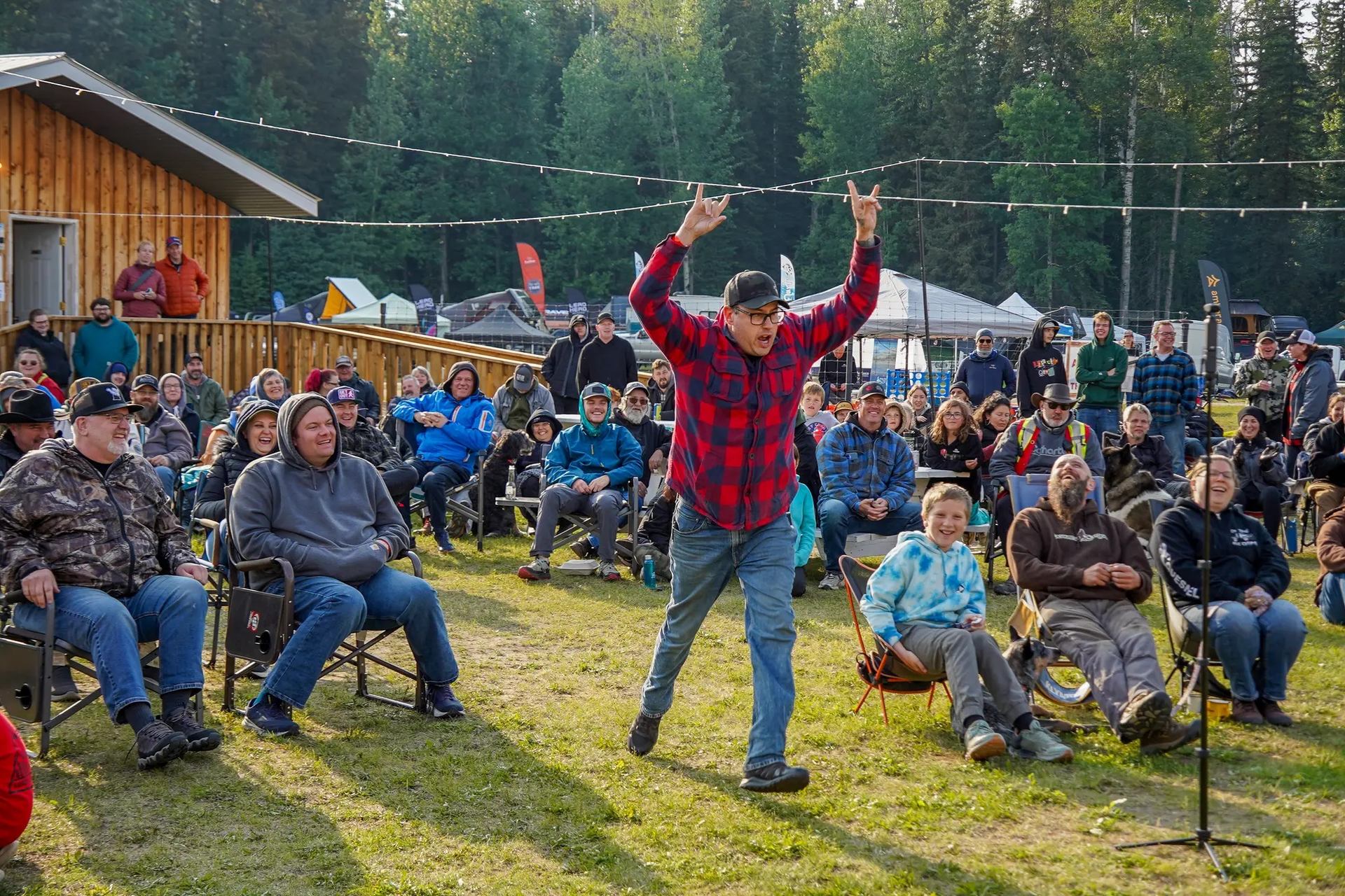 An excited man in a red plaid shirt raises his arms in "rock on" gestures before a large outdoor audience seated on a grassy lawn.