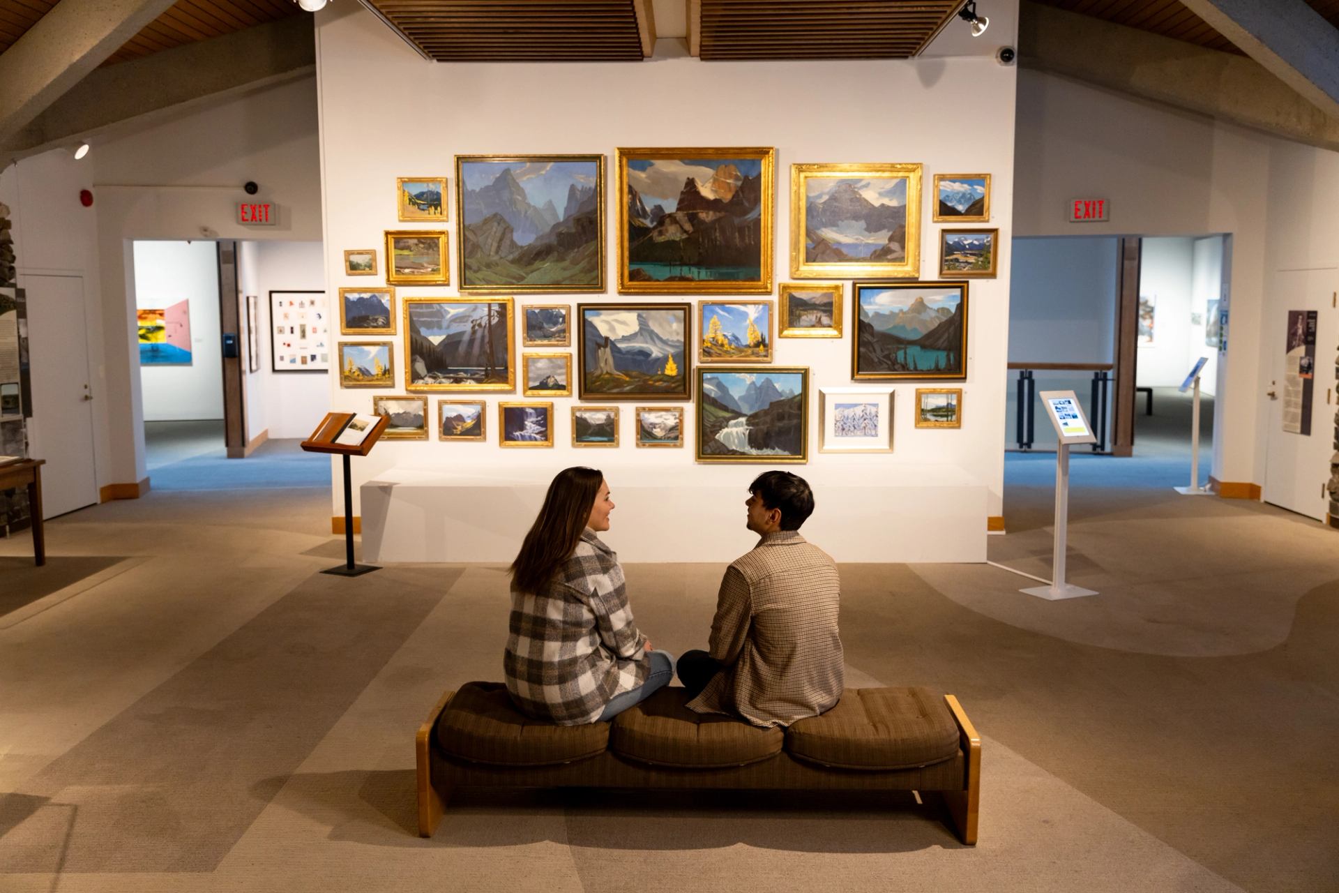 Two people sit on a bench facing a gallery wall of framed mountain paintings.