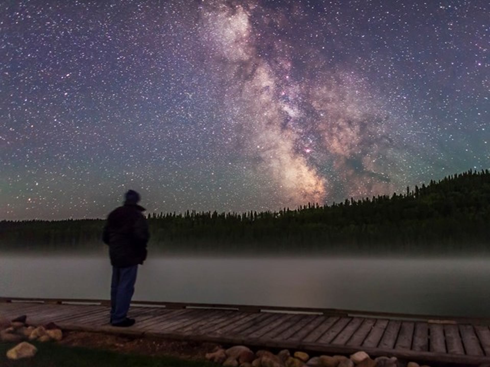 A man stands on a dock looking at the night sky across an alpine lake.