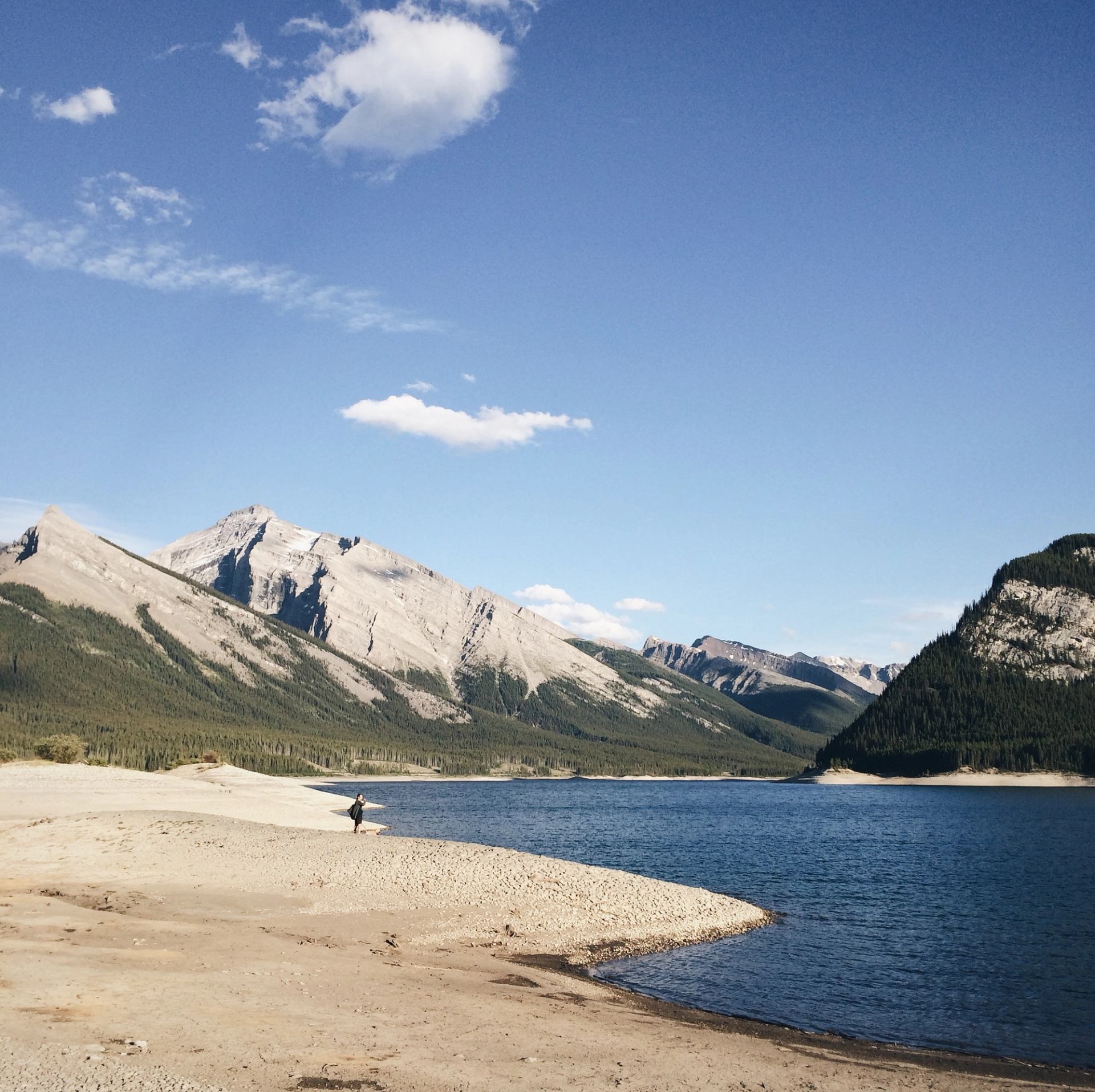 Rocky mountain lake with sandy shoreline, distant hiker, and clear blue sky.