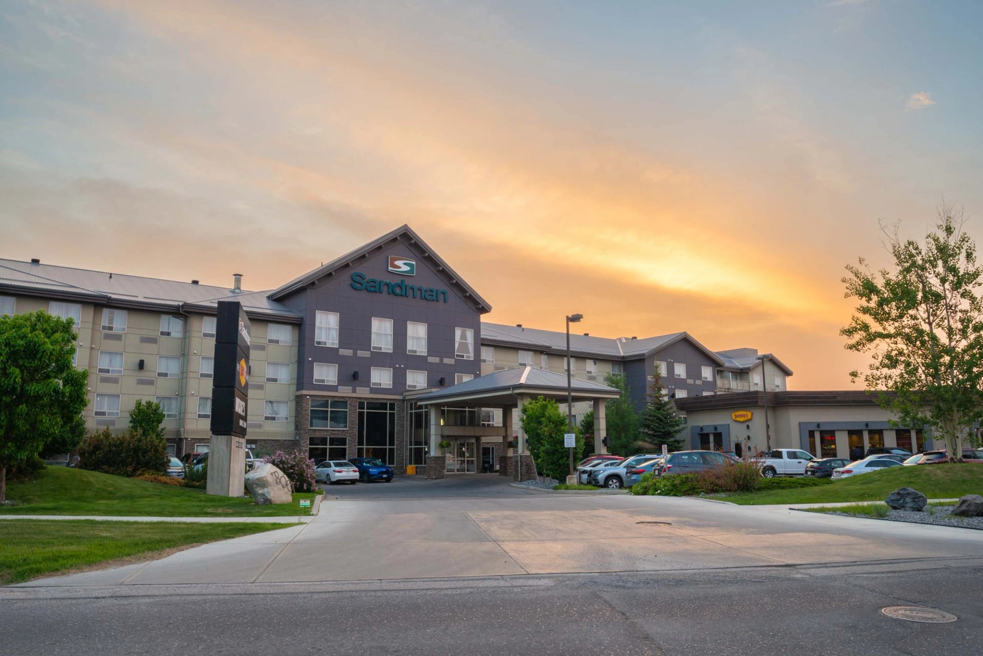 Sandman hotel exterior with parking lot, cars and trees under an orange sky.