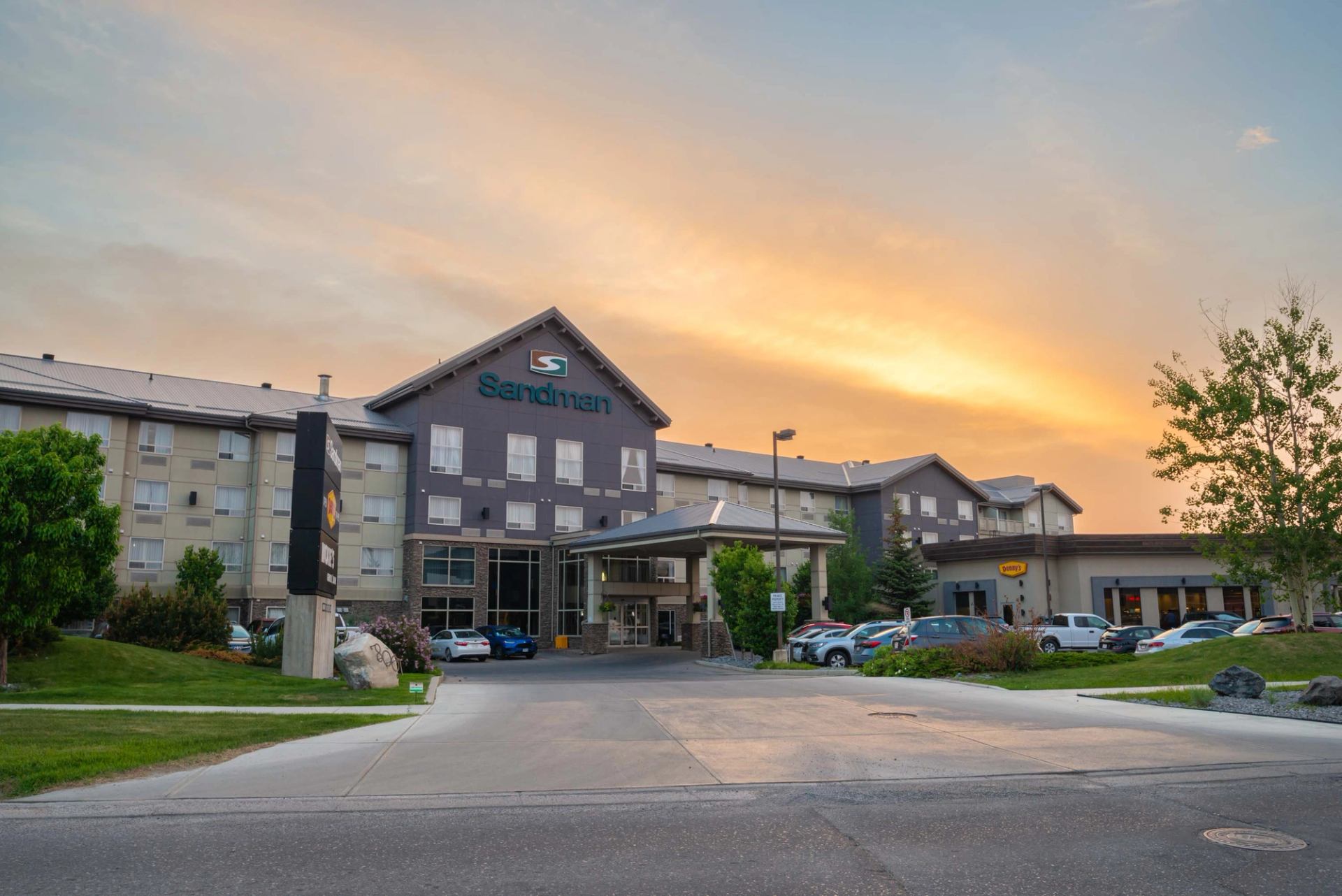 Sandman hotel exterior with parking lot, cars and trees under an orange sky.
