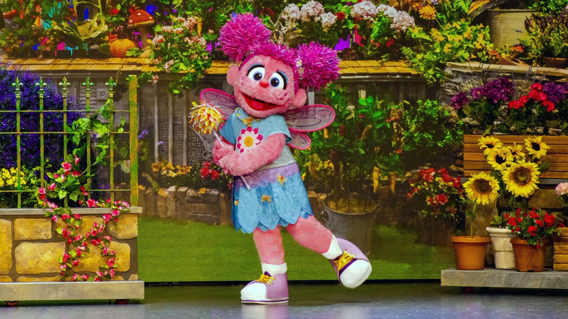 Pink fairy puppet with wings and wand posing onstage before colourful garden backdrop.