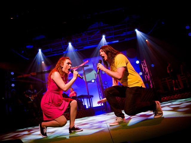 Two performers singing on stage with colorful lights and musical gear in the background.