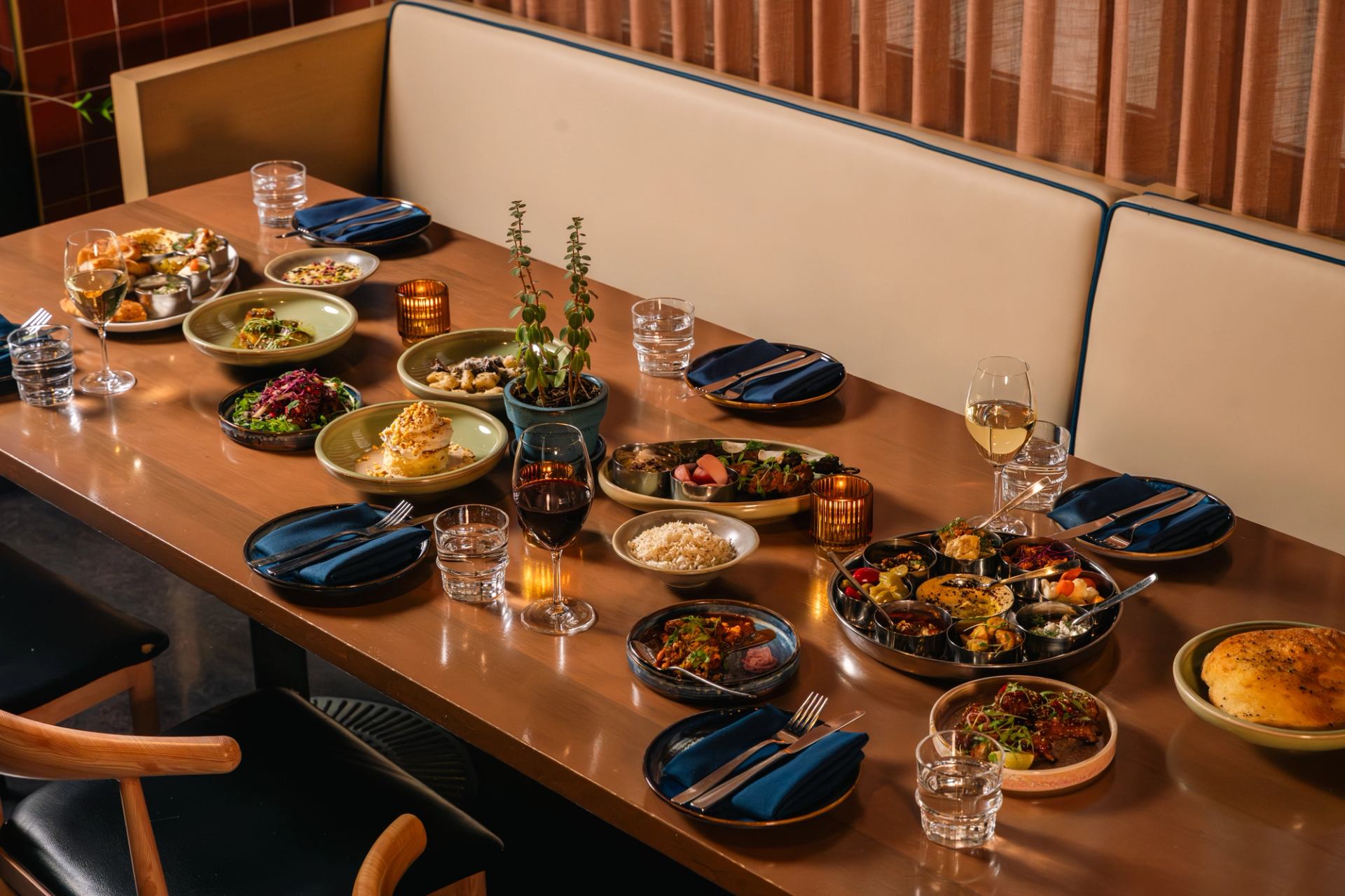 Restaurant table with assorted shared dishes, wine glasses, candles and potted plants.