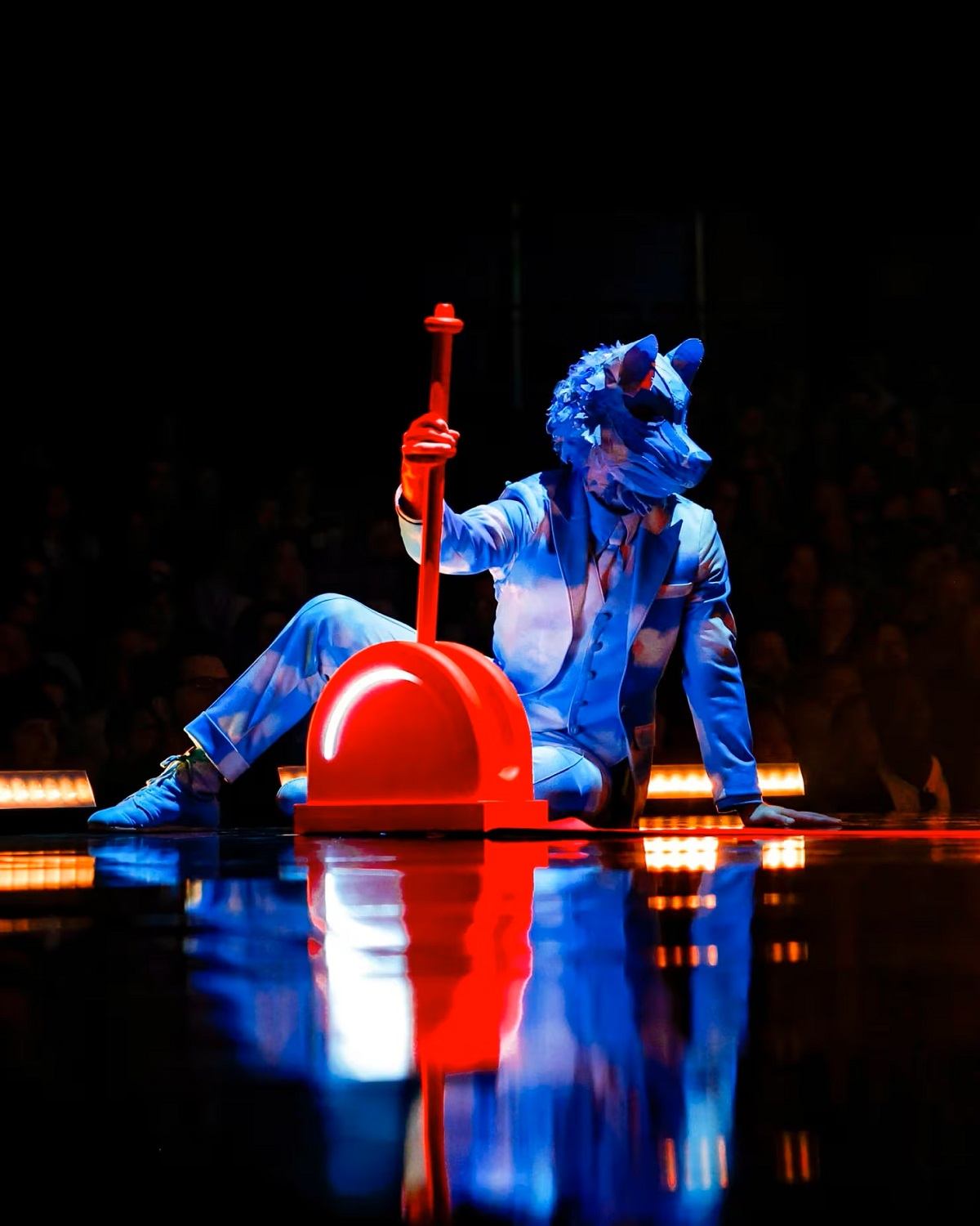 Performer in blue costume holds red spear-like prop on dark stage with audience in background.