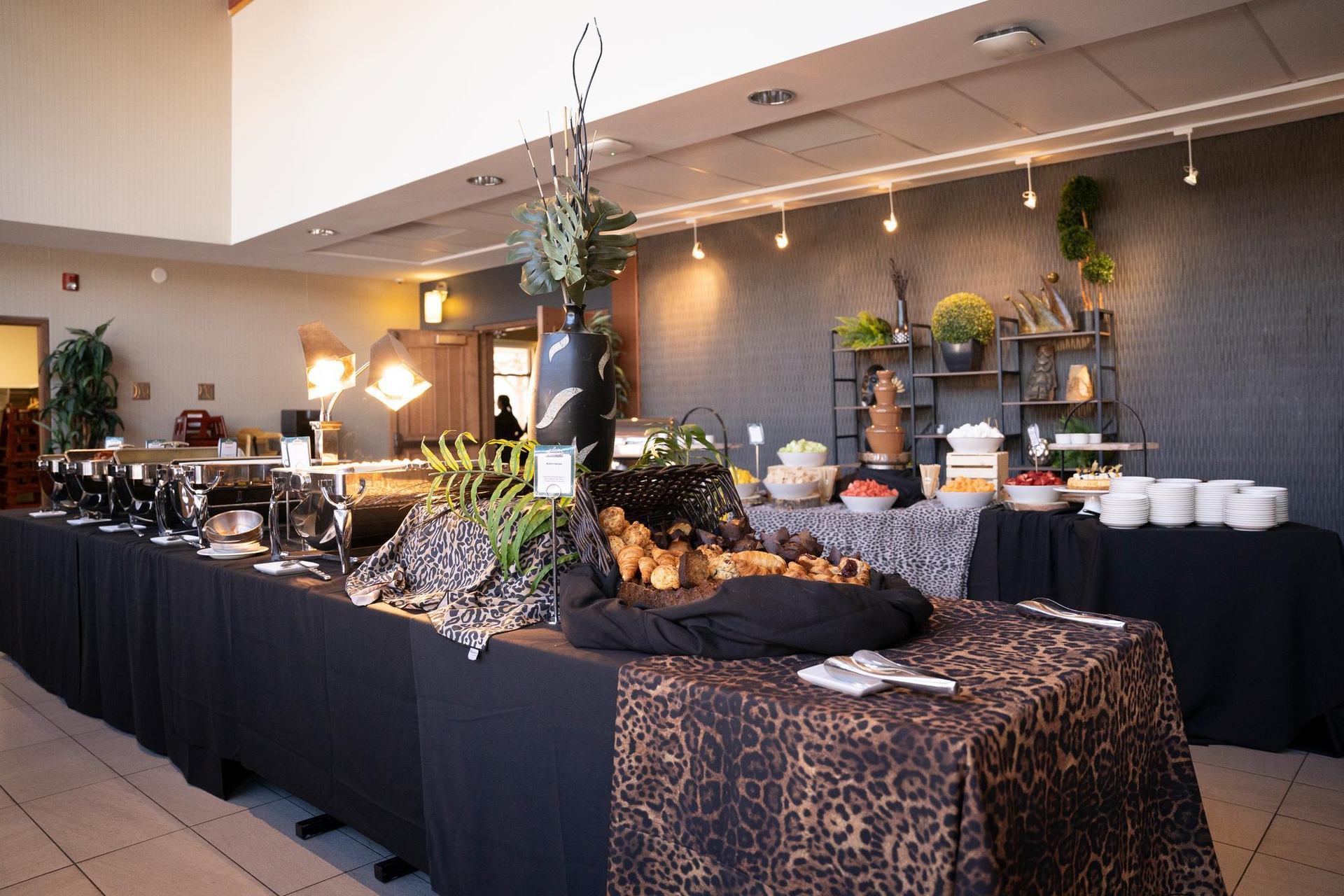 A long buffet table is laden with hot dishes, pastries, fruits, and a chocolate fountain, featuring black and leopard print linens.
