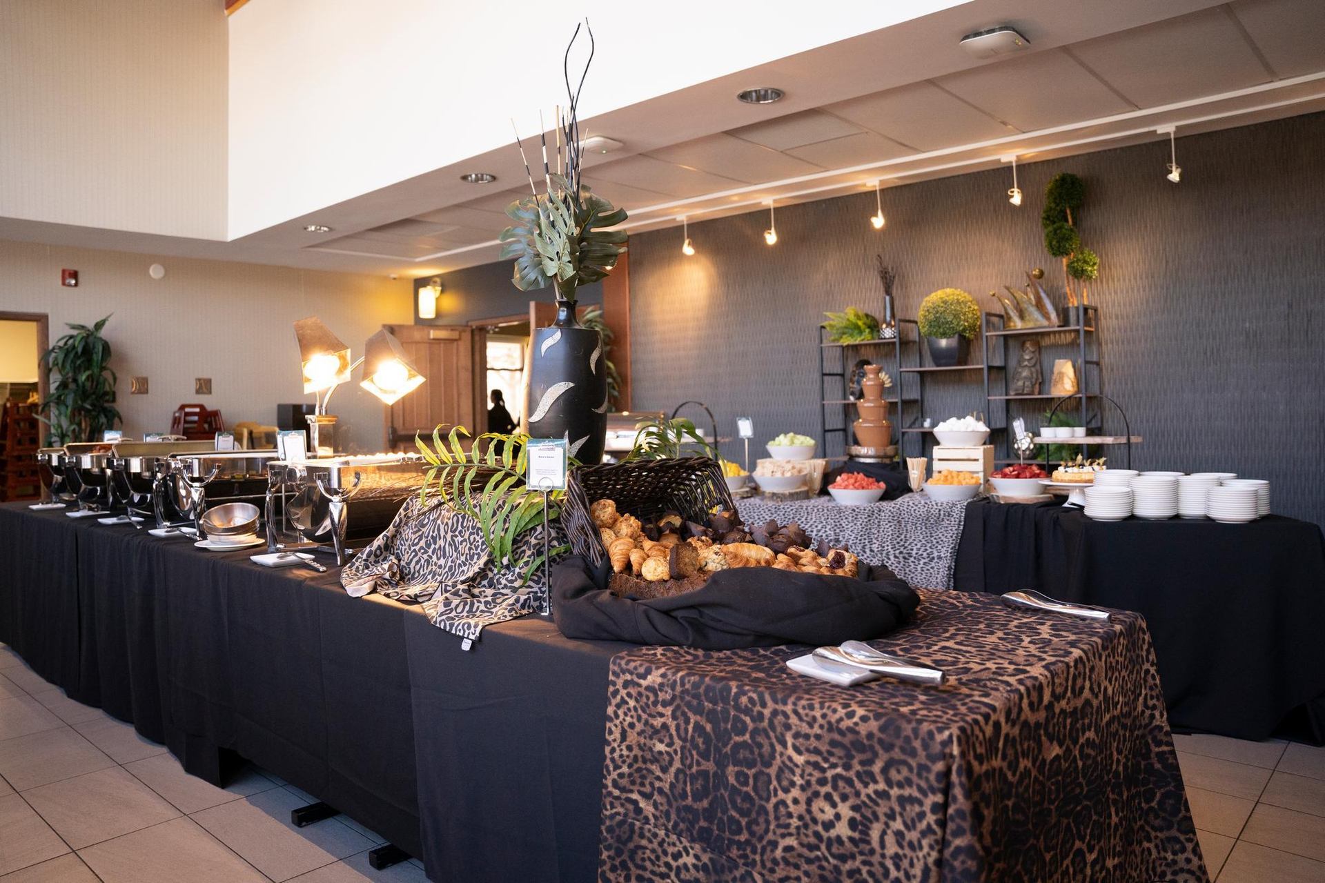 A long buffet table is laden with hot dishes, pastries, fruits, and a chocolate fountain, featuring black and leopard print linens.