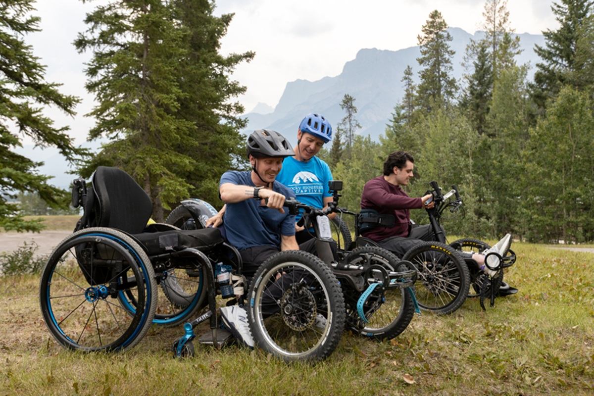 Three cyclists in adaptive off-road wheelchairs pause on a grassy trail among evergreen trees.