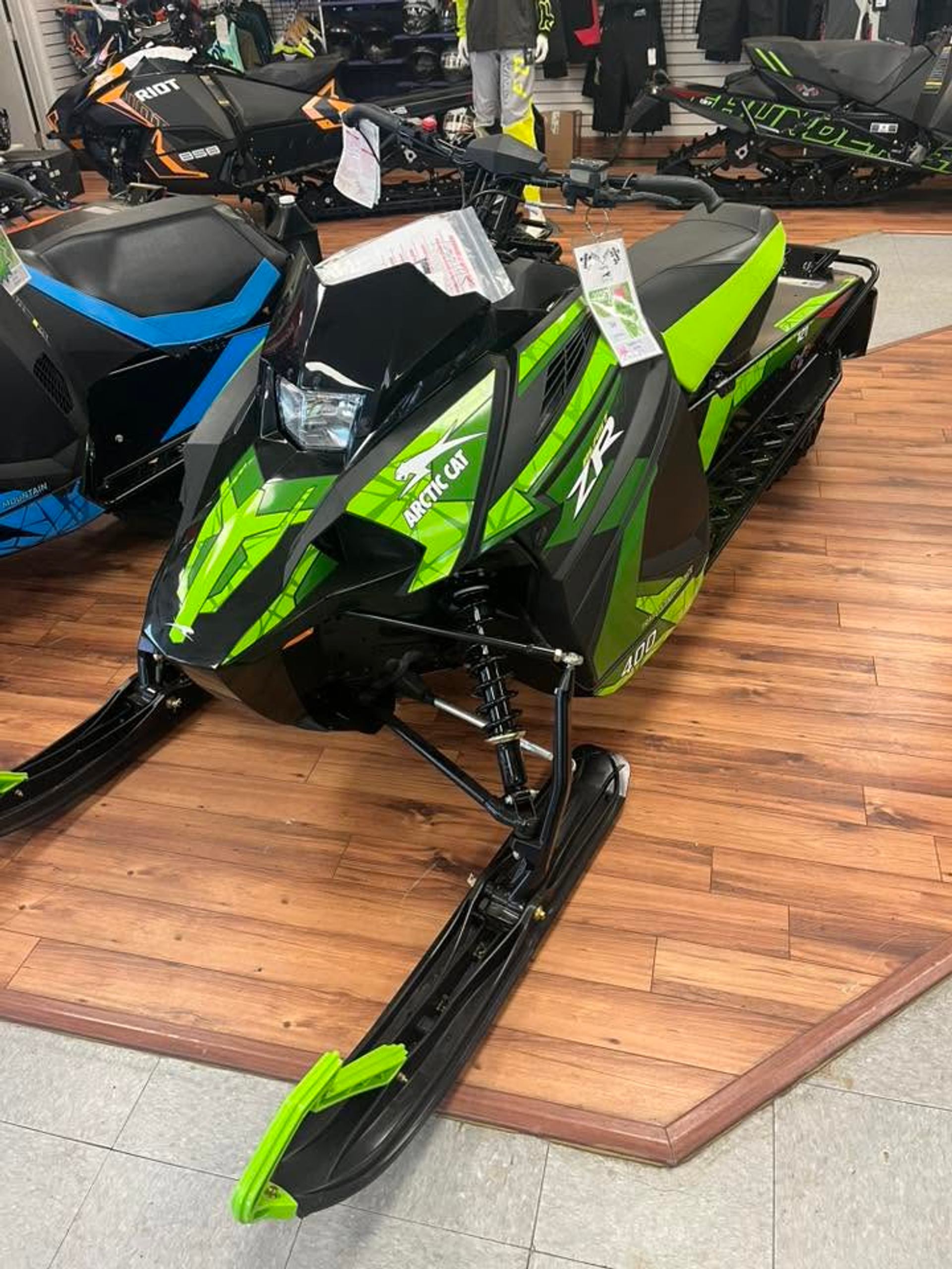 Green and black Arctic Cat snowmobile displayed on a wood floor among other snowmobiles.