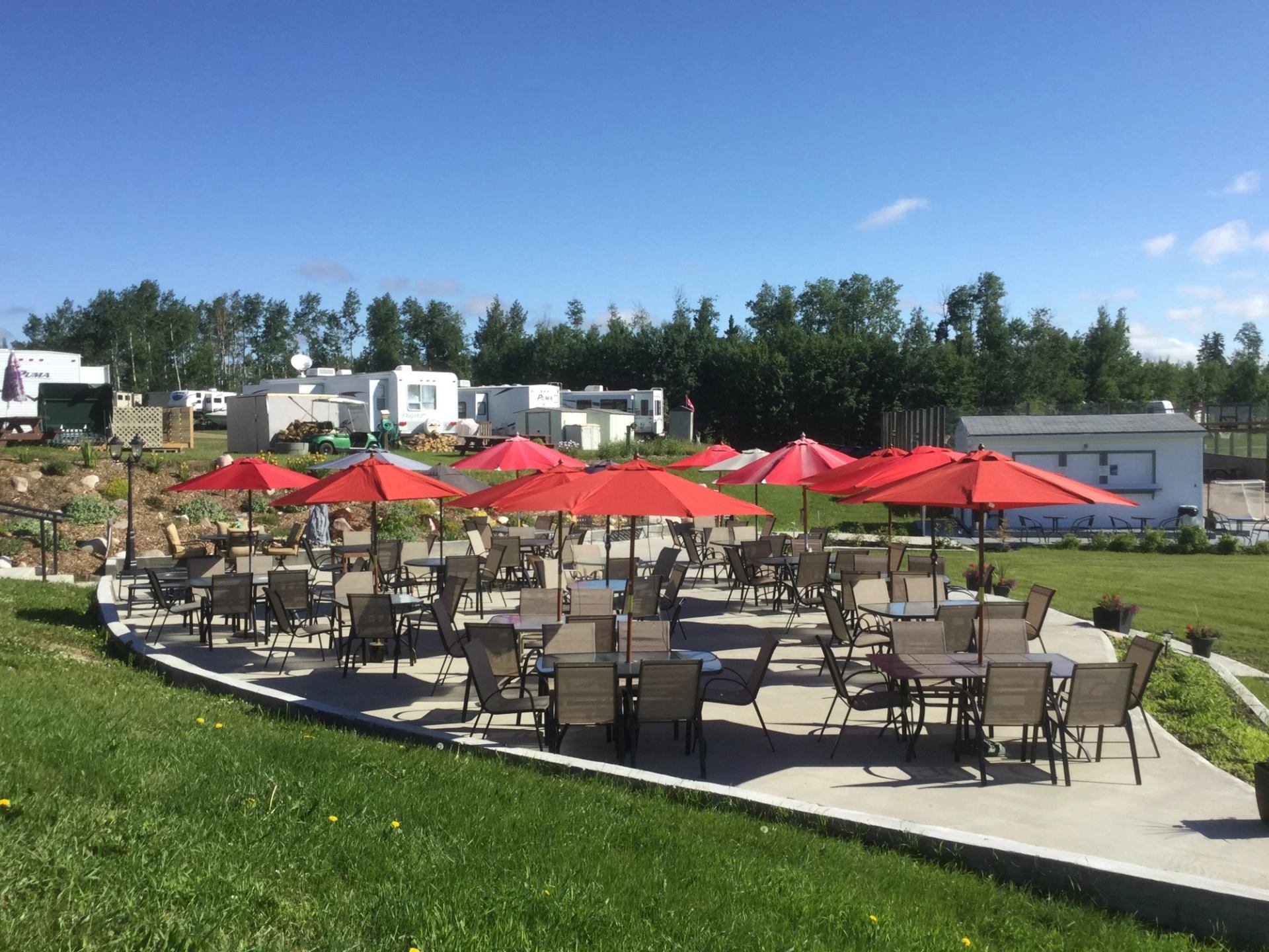 Outdoor tables with red umbrellas on patio surrounded by grass and trees.