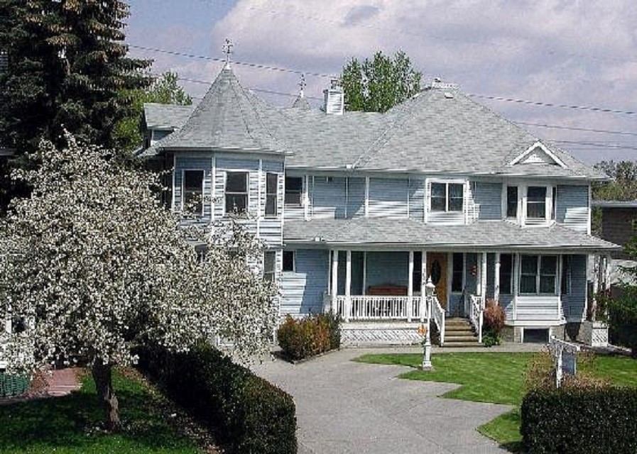 Large Victorian-style house with a gray roof, white siding, and blooming trees in front yard.