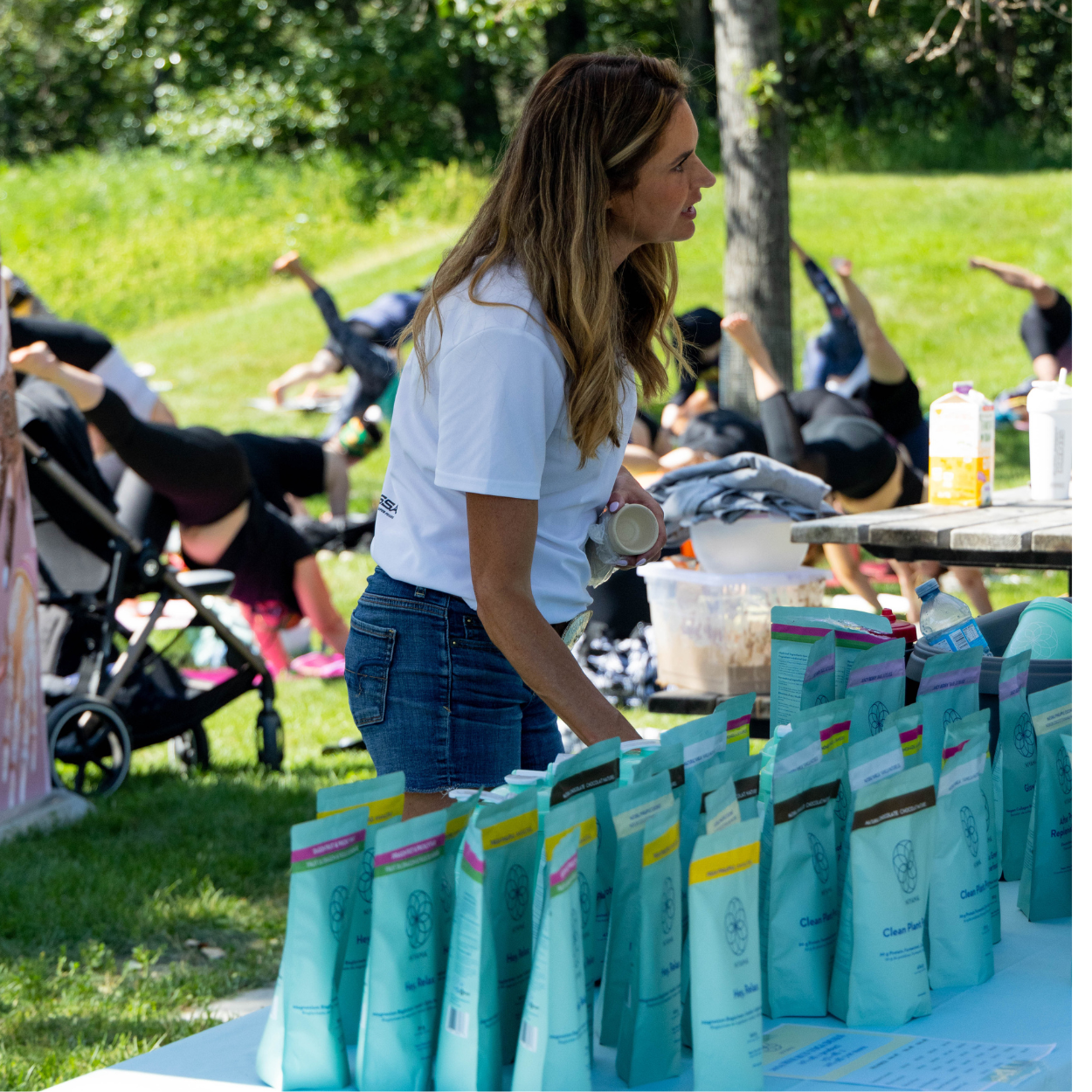 Volunteer stands at a table of wellness products while participants practice yoga on mats in a park.