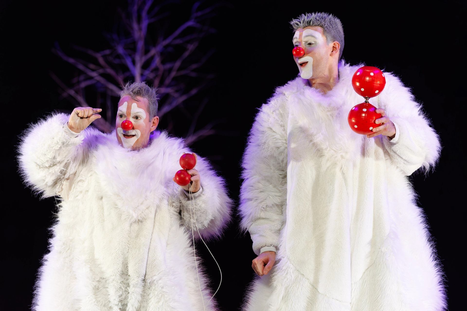 Two clowns in fluffy white costumes hold red juggling props on a dark stage.