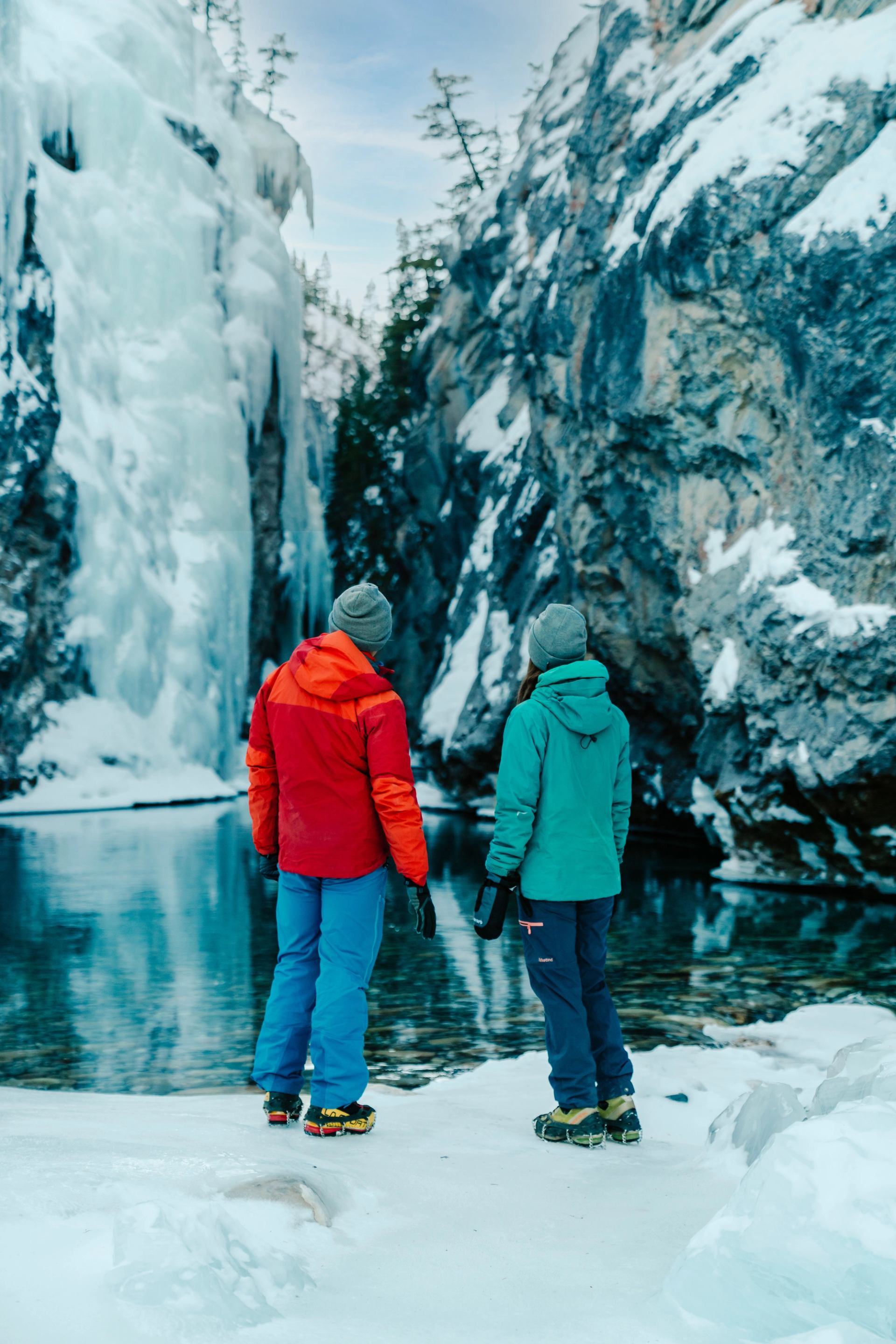 Two people in winter gear stand by a clear river, facing an icy waterfall and snow-covered cliffs.