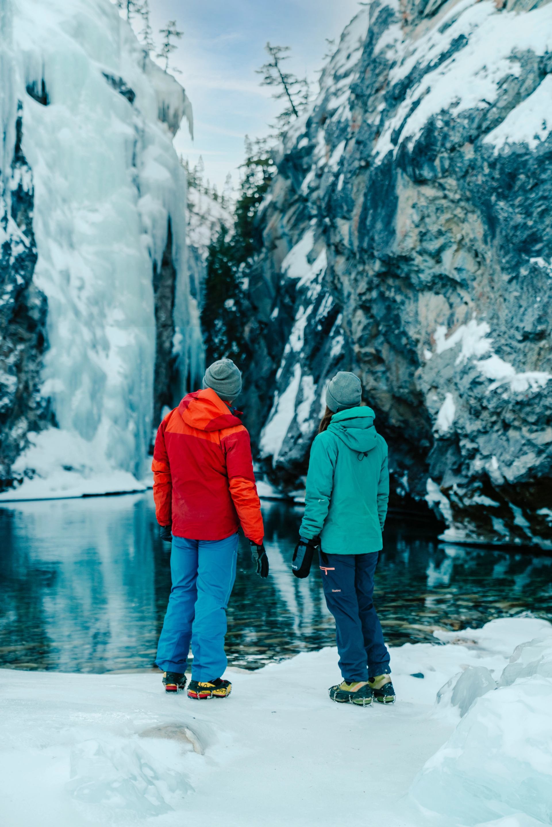 Two people in winter gear stand by a clear river, facing an icy waterfall and snow-covered cliffs.