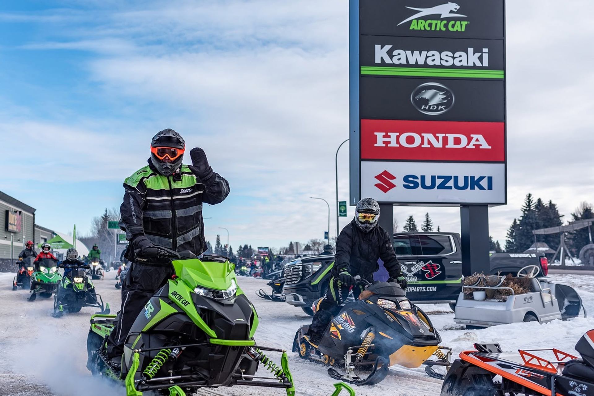 Snowmobilers gather beside Arctic Cat, Kawasaki, Honda and Suzuki dealership signs.